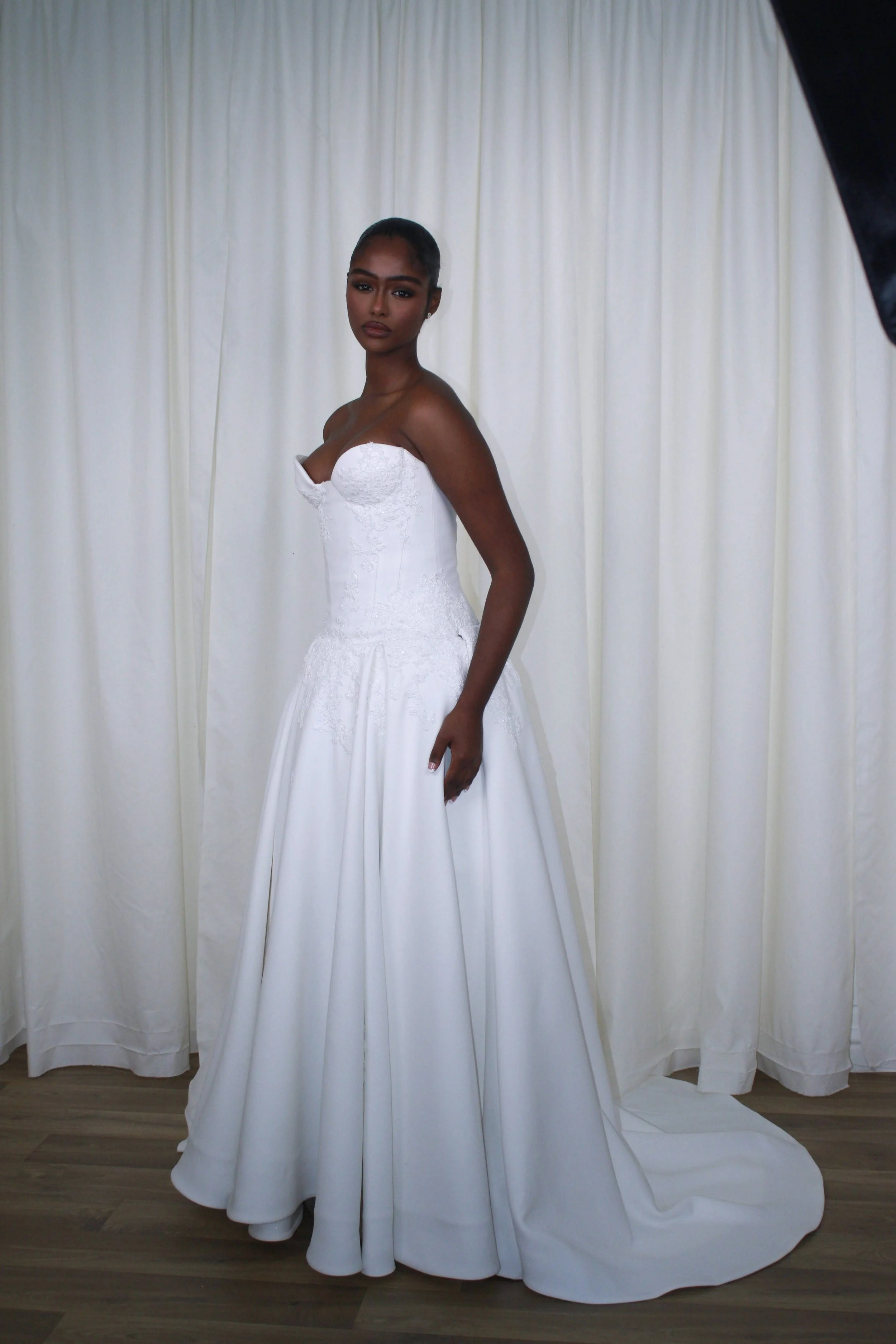 A woman in a strapless white wedding gown with lace detailing, standing on a wooden floor in front of white curtains.