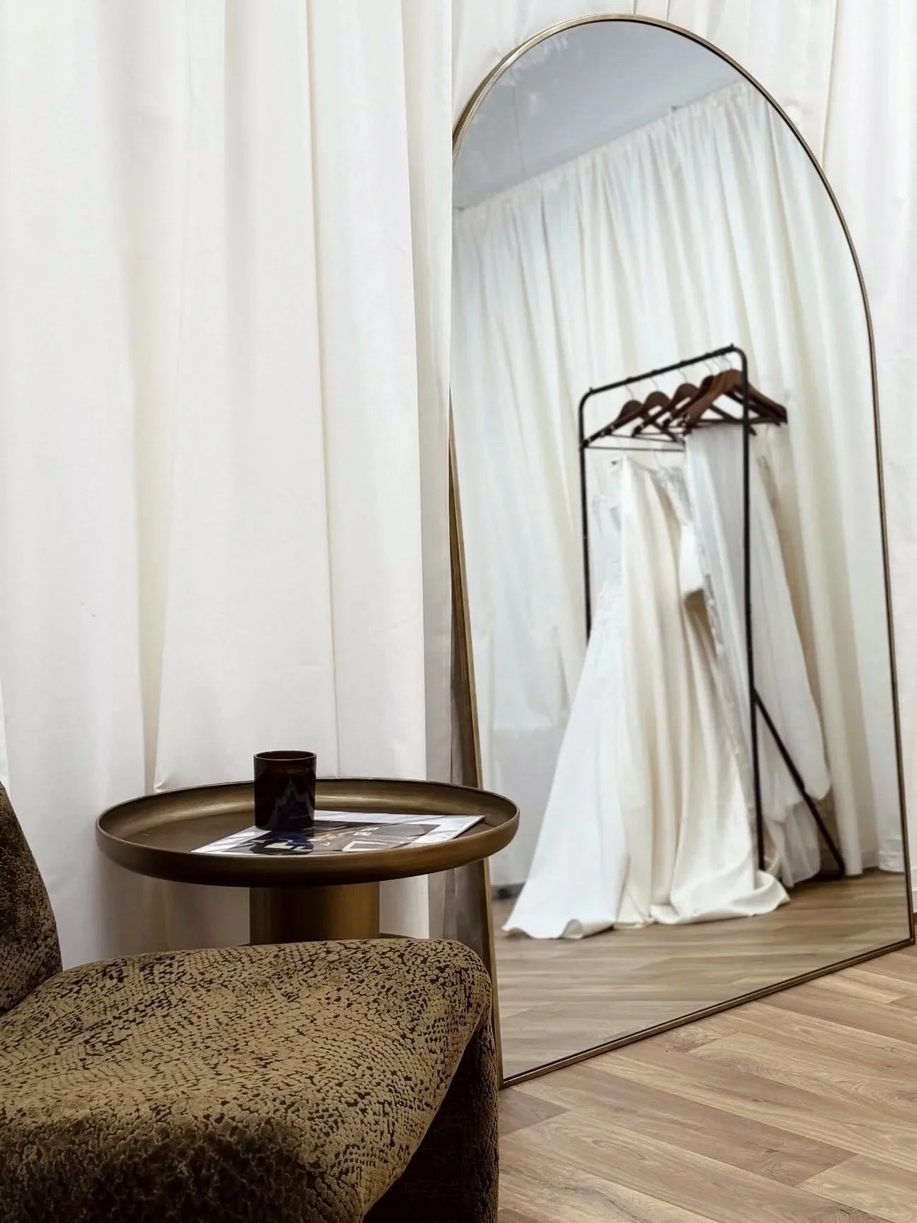 A fitting room with a large oval mirror, a dress on a hanger rack, a brown round table with a magazine and cup, and a plush patterned chair, all surrounded by beige curtains.