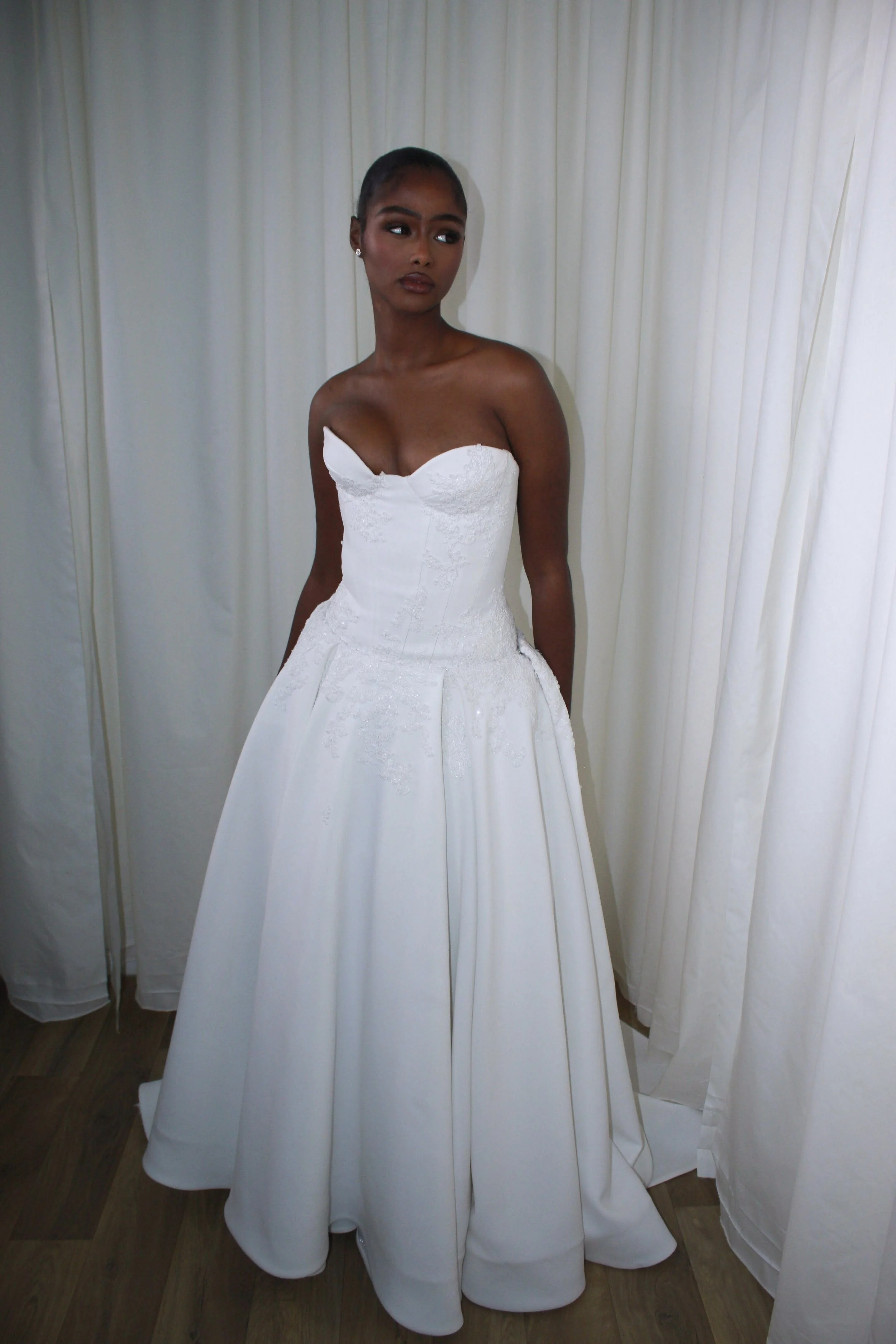A woman in a strapless white wedding dress standing in front of white curtains.