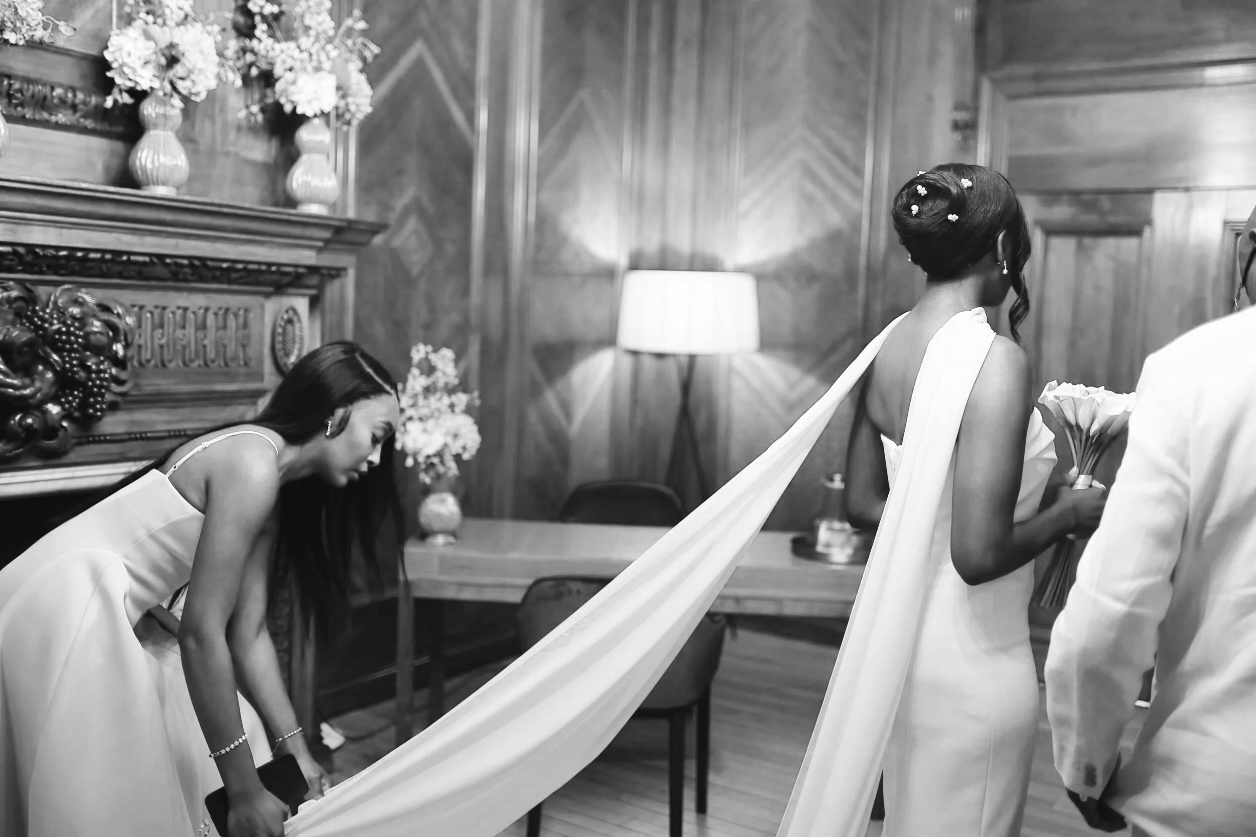 A bride in a wedding dress holding a bouquet is being assisted by a woman in a white dress, with another woman leaning forward nearby, inside a wood-paneled room with floral arrangements and a table.