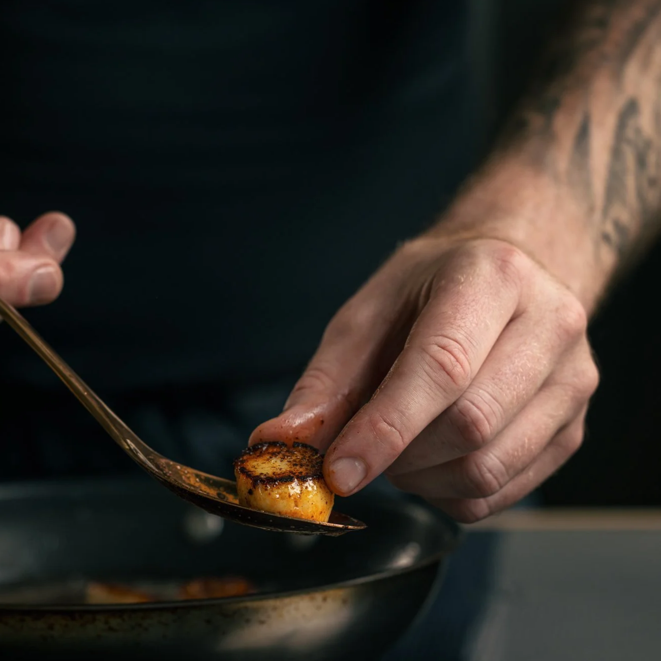 Close-up of a person's hand placing a grilled scallop on a plate with dark sauce.