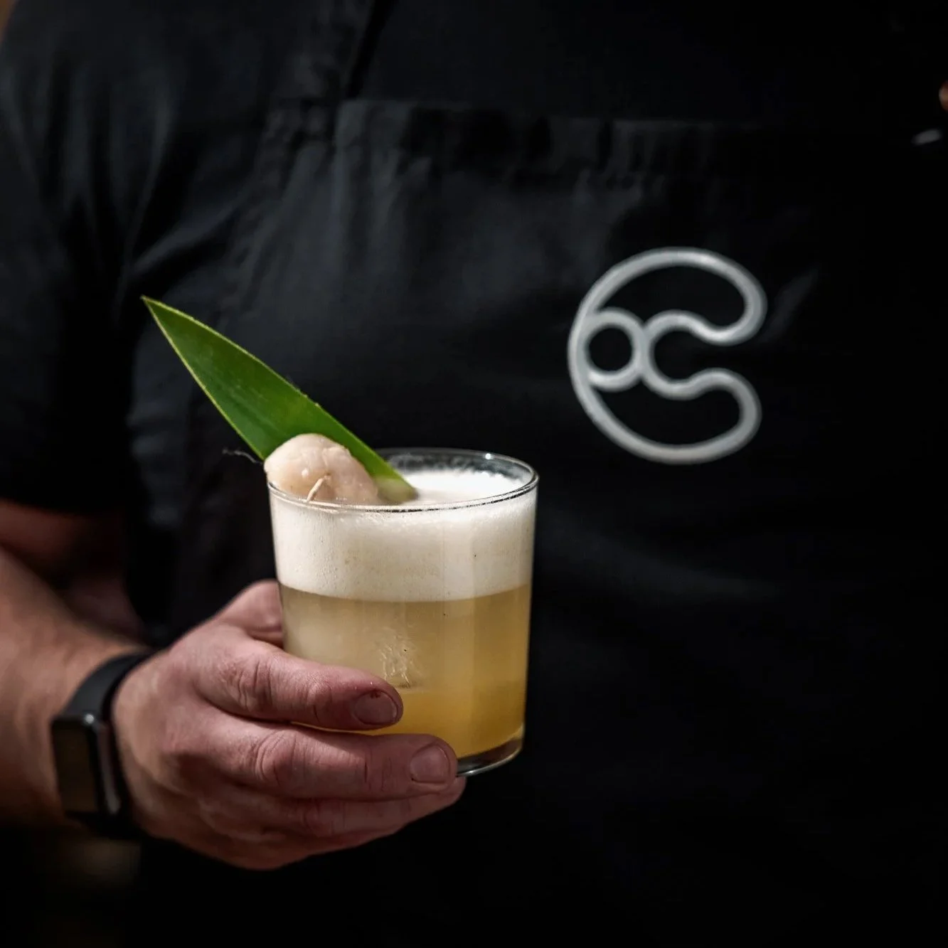 Person holding a cocktail garnished with a pineapple leaf and a piece of lychee, with foam on top, in a clear glass.