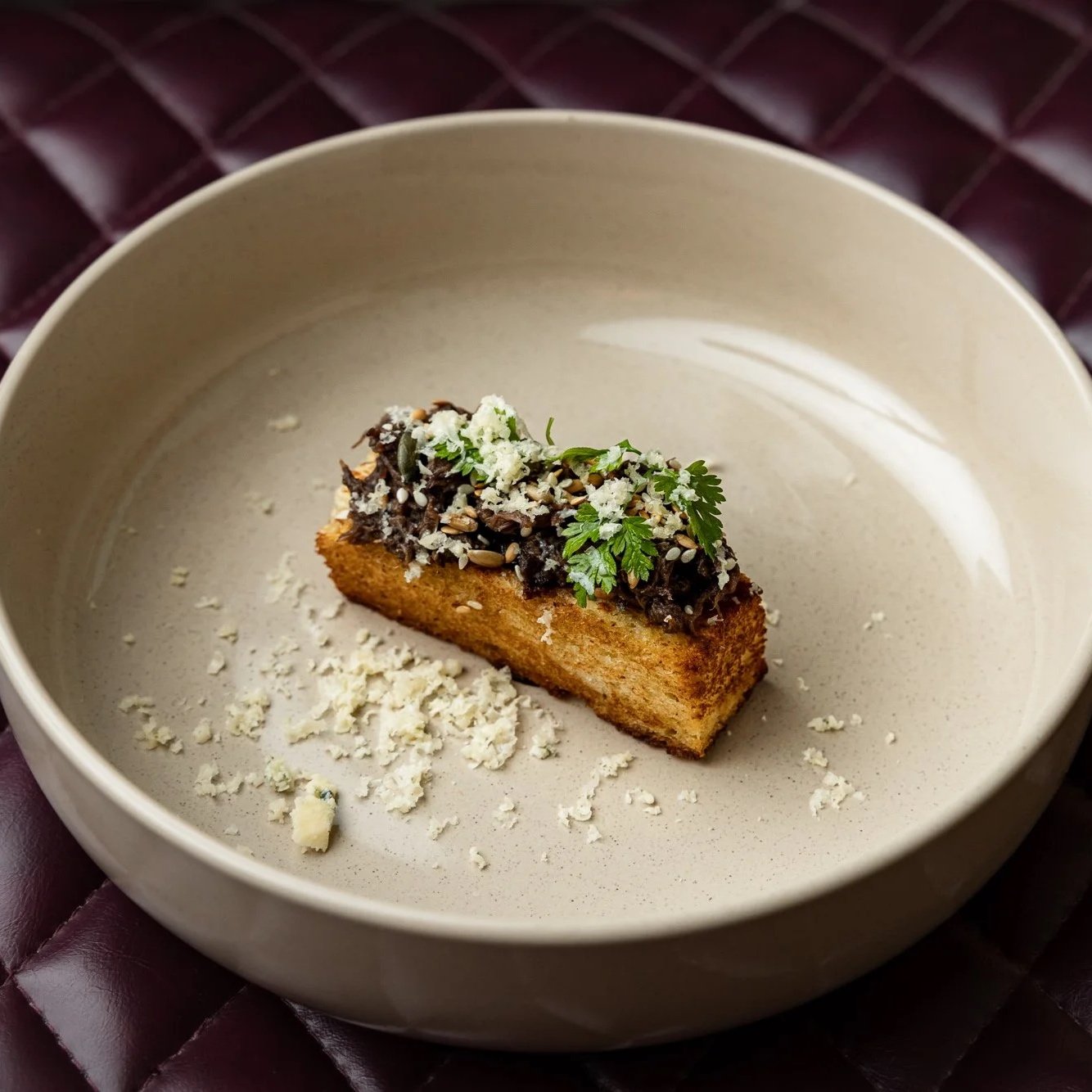 An artfully plated dessert featuring a rectangular piece of cake topped with chocolate, sunflower seeds, fresh herbs, and grated white chocolate, served in a beige bowl on a dark quilted surface.
