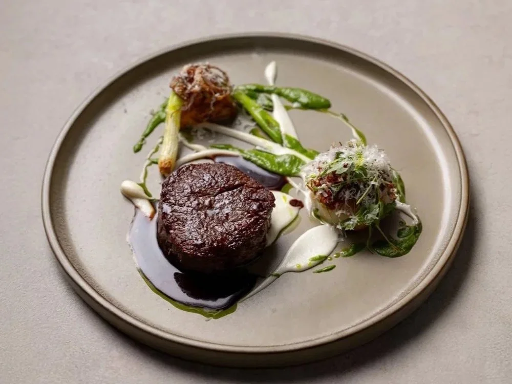 A gourmet plate featuring a seared piece of steak topped with a dark sauce, accompanied by a small stack of green vegetables, a small round salad topped with shredded cheese, and garnished with sauces and sprouts.