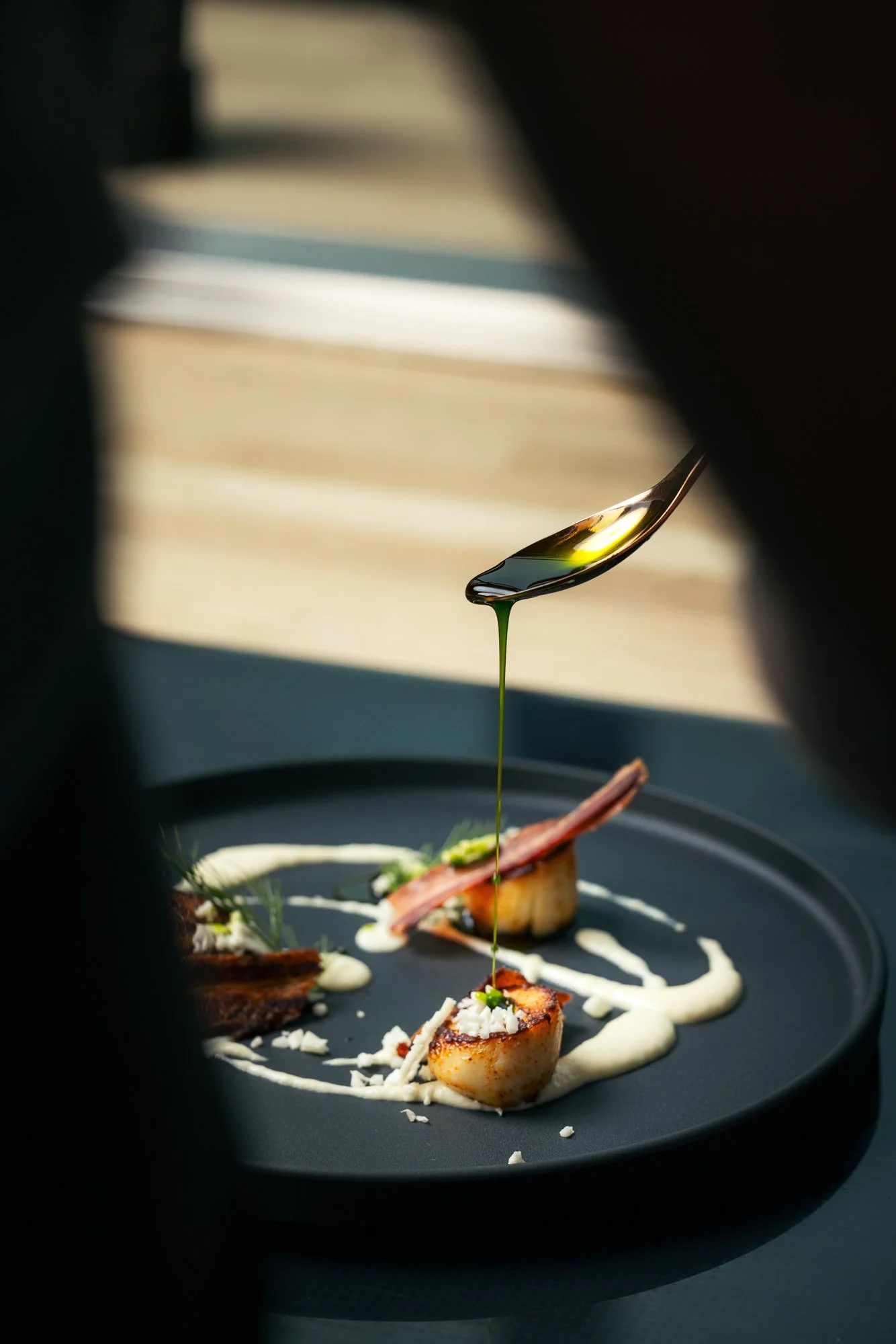 A close-up of a gourmet dish with scallops, bacon, and sauce on a black plate, with olive oil being drizzled from a spoon.
