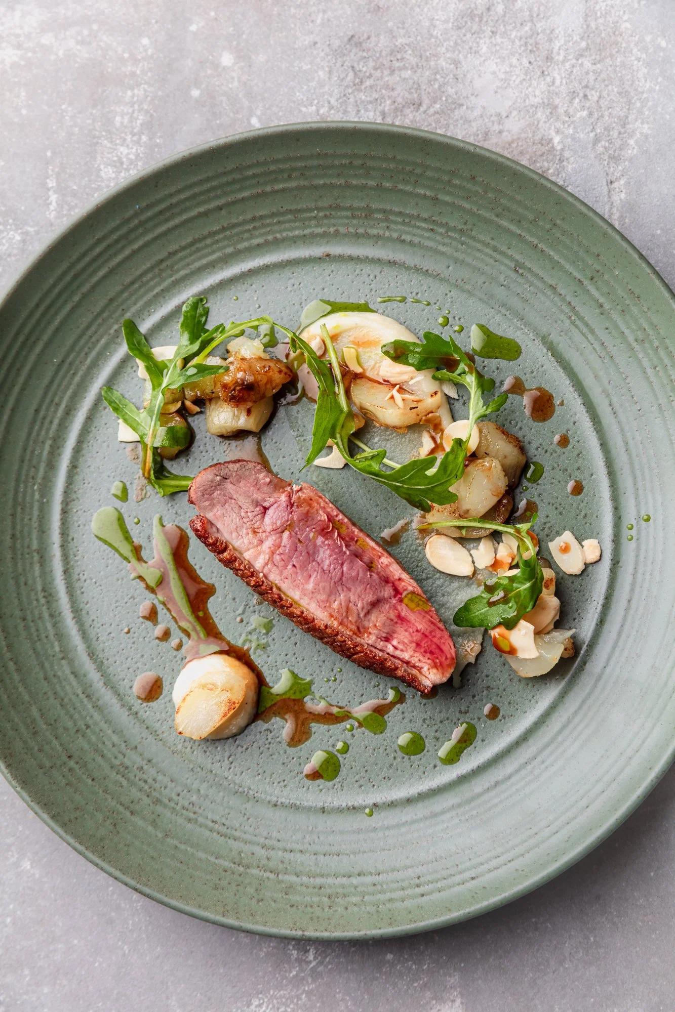 A gourmet dish on a green plate featuring a slice of medium-rare steak, garnished with greens, roasted garlic, and a drizzle of sauce.