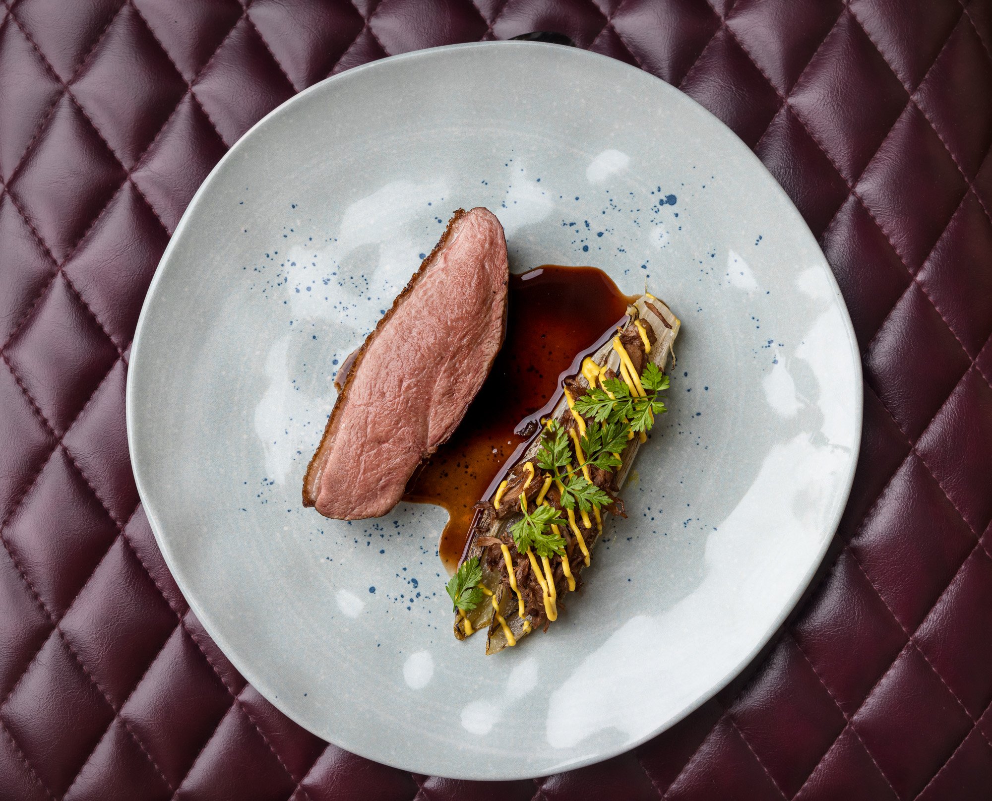 A gourmet dish featuring a slice of cooked meat and a vegetable topped with sauce and garnished with cilantro, served on a white plate.