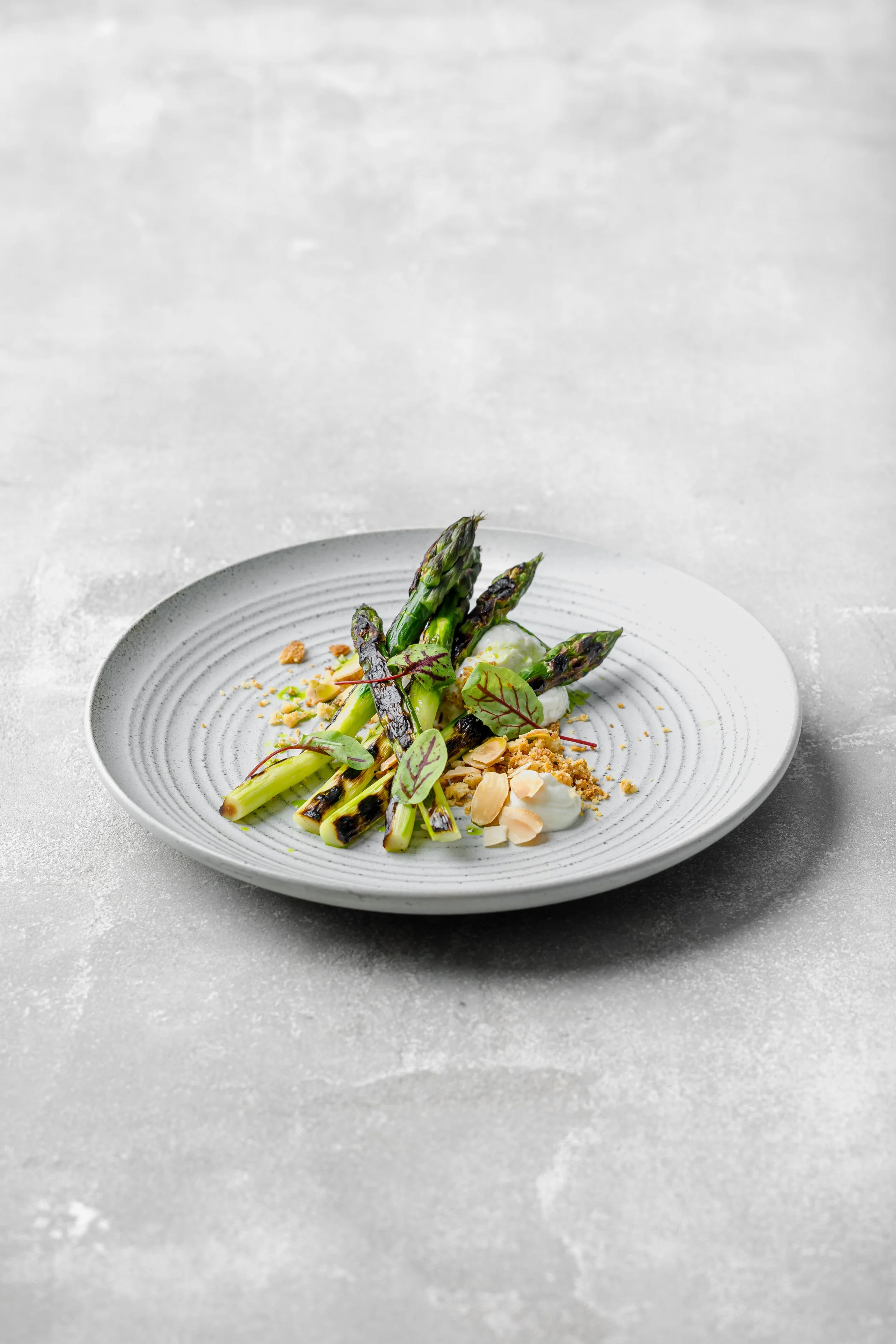 A white plate with grilled asparagus spears, garnished with microgreens, slivered almonds, dollops of white sauce, and crumbles, set on a textured light gray surface.