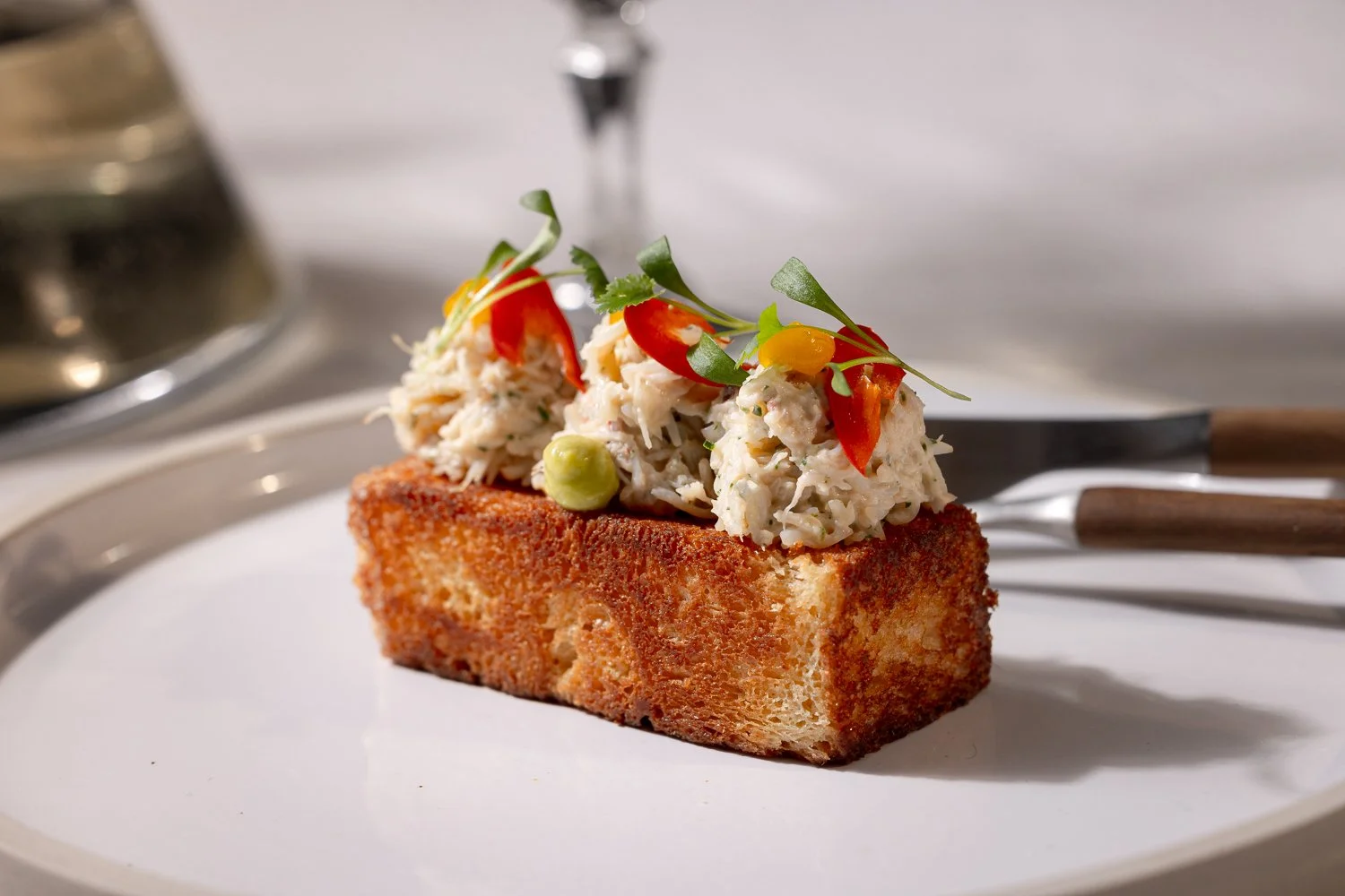 A slice of toasted bread topped with crab salad, garnished with cherry tomato slices, microgreens, and green peas, served on a white plate.