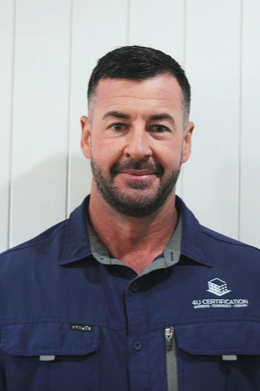 A man with short dark hair and a beard, wearing a navy blue work shirt with a logo that says "4U CERTIFICATION" and a smaller tag that says "SYNER" in front of a white panel wall.