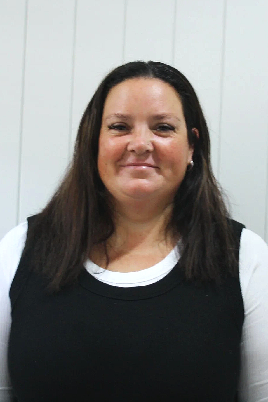 A woman with shoulder-length dark hair, smiling, wearing a black top with white sleeves, standing against a white wood-paneled wall.