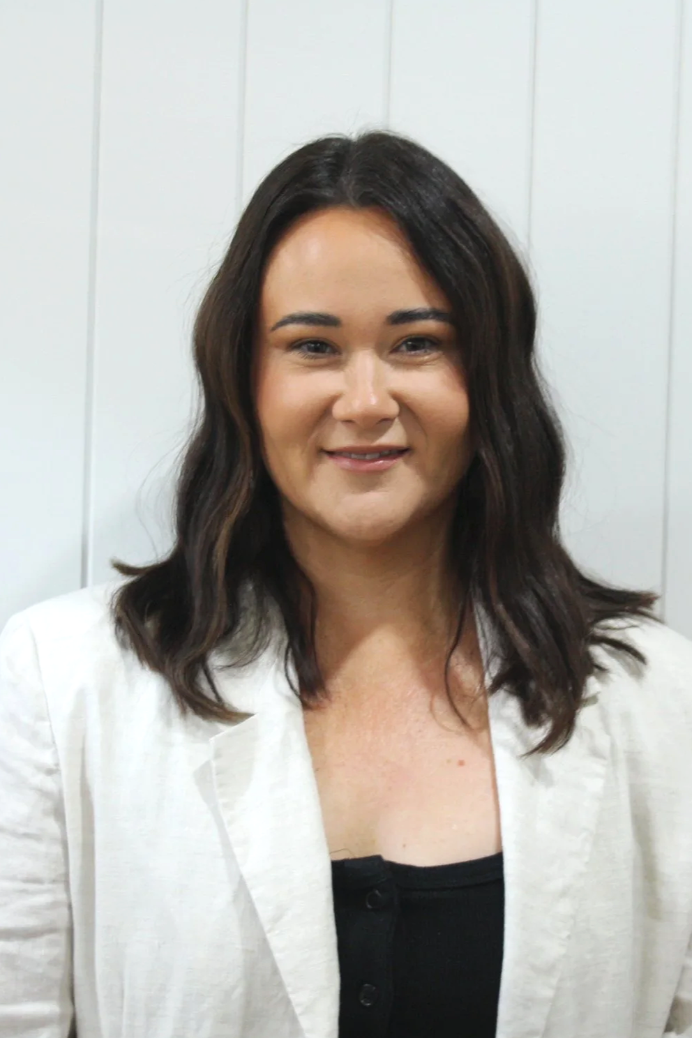 A woman with shoulder-length wavy dark hair, wearing a white blazer over a black top, smiling at the camera in front of a white paneled wall.