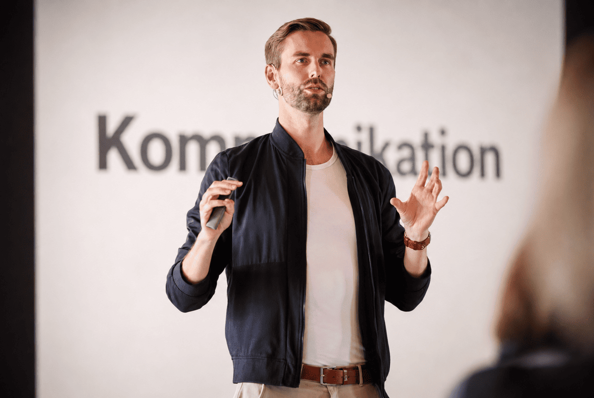 Vortragende Person spricht vor Publikum über Kommunikation und unterstützt die Aussagen mit Gestik. Das Bild verdeutlicht die Bedeutung von Körpersprache in einer politischen Rede.