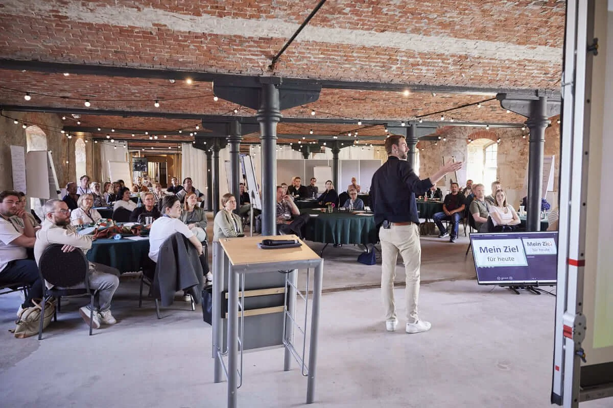 Ich (ein Mann in den 30ern) halte einen Vortrag. Das Publikum guckt auf die Leinwand, auf der gerade angezeigt wird "Mein Ziel für heute".