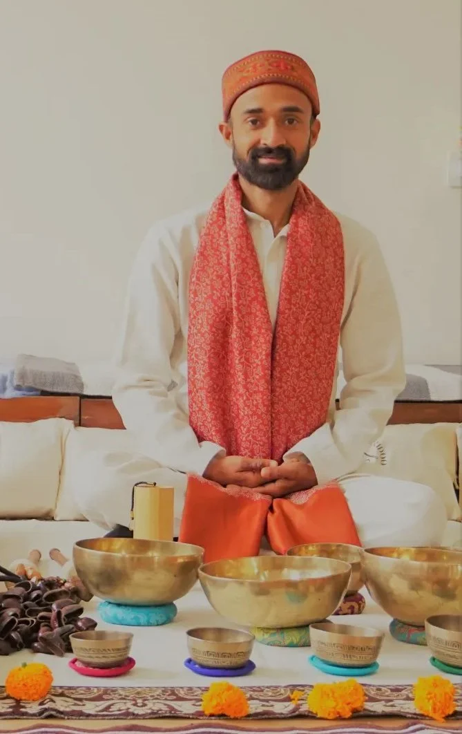 Man in traditional Indian attire sitting cross-legged with prayer beads, surrounded by brass bowls, maracas, and marigold flowers, performing a ceremonial ritual.