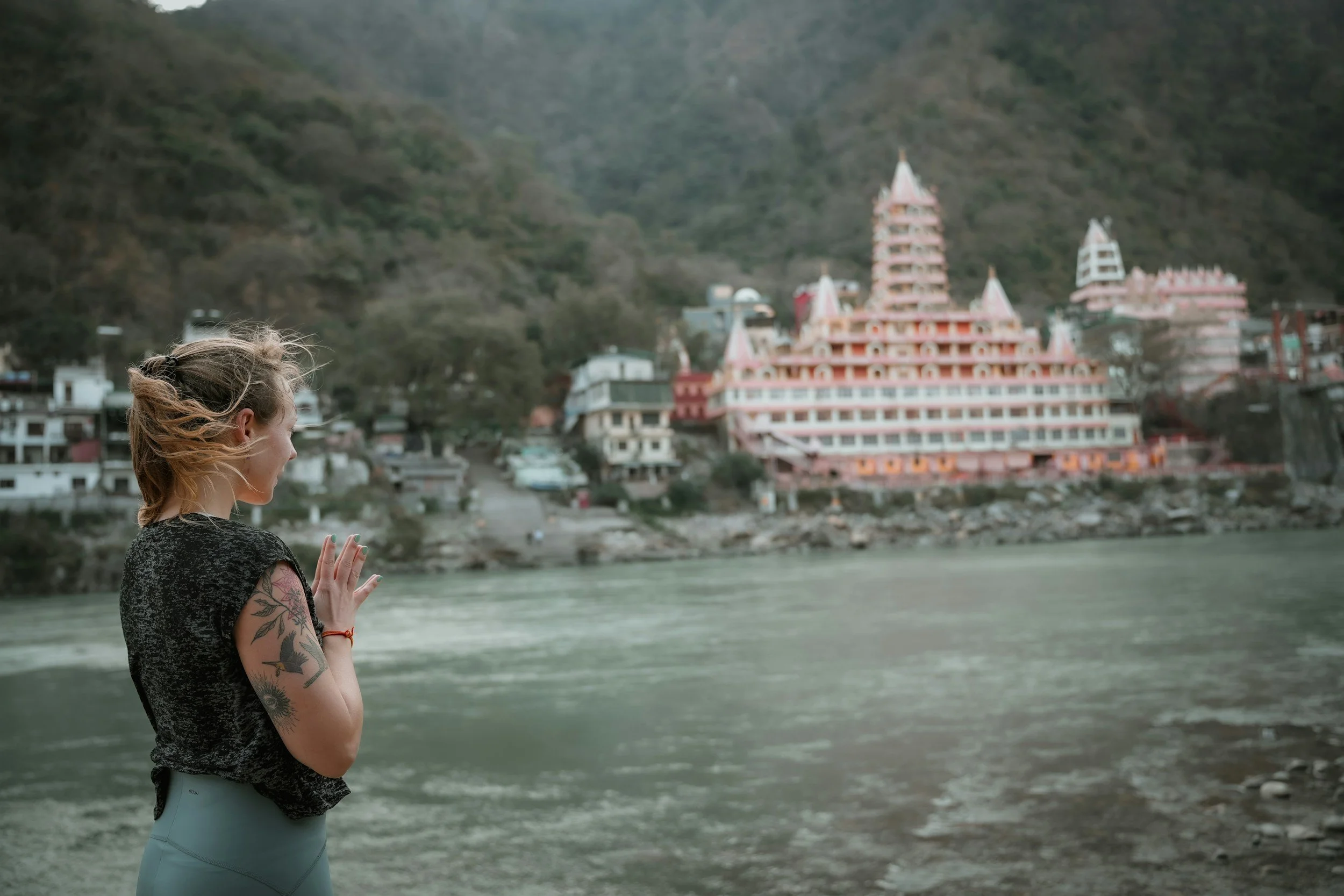 A woman with tattoos on her arm standing by a river with a large, pink temple on a hill in the background.