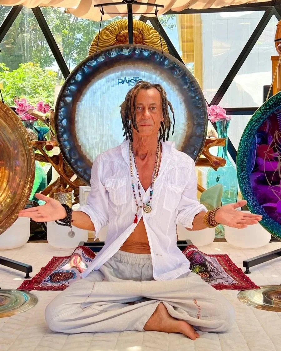 A man with dreadlocks sits cross-legged on a mat, meditating or practicing yoga indoors. He is wearing a white shirt open to his chest, beige pants, and various necklaces and bracelets. Behind him are large, colorful metal or glass decorative objects, including a large circular piece with sparkling accents. The setting is bright and airy, with sunlight streaming through large windows and plants visible outside.