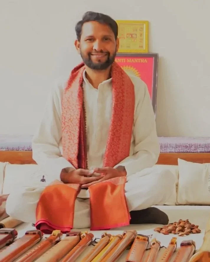 A man with a beard and mustache, smiling, is sitting cross-legged on a cushion. He is wearing traditional Indian clothing, a white kurta and orange shawl. In front of him, there are musical instruments. The background shows a wall with framed posters or certificates.