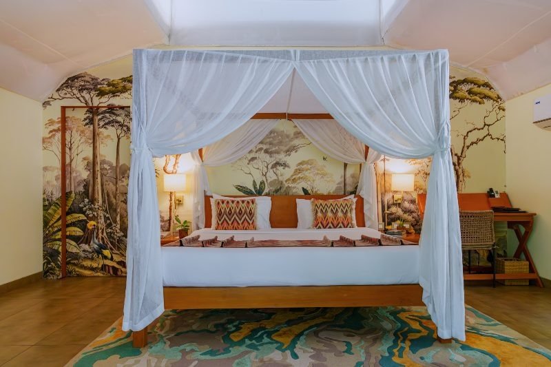 A bedroom with a four-poster bed fitted with white linens and patterned throw pillows, featuring a sheer white canopy. The room has a nature-themed mural on the wall behind the bed, depicting trees and foliage. There are bedside lamps, a desk with a wicker chair, and a colorful area rug on the wooden floor.