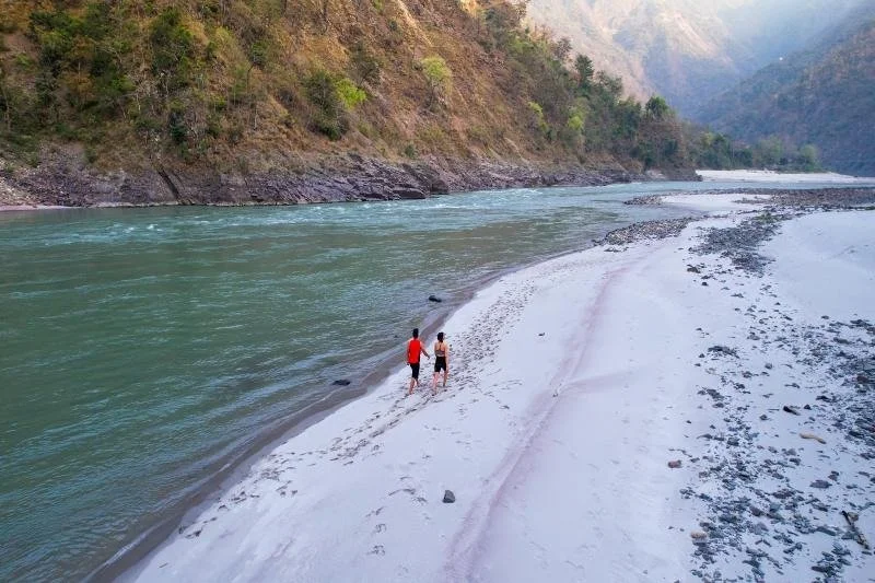 Two people walking along a sandy beach beside a wide, calm river with greenish water, surrounded by forested mountains.