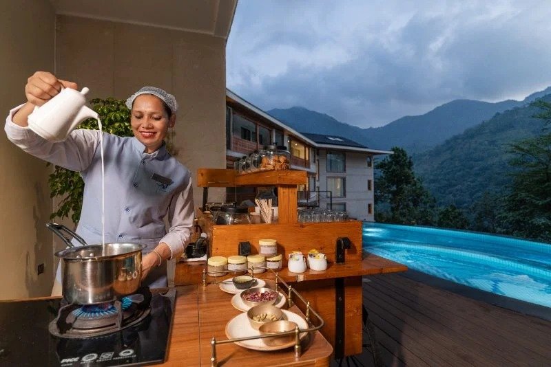 A woman in chef attire pouring milk into a saucepan while cooking on a stove in an outdoor kitchen with mountains in the background.