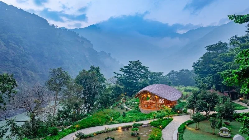 Landscape view of a mountain valley with lush greenery, trees, and misty mountains in the background. There is a curved pathway leading to a wooden pavilion with a thatched roof.