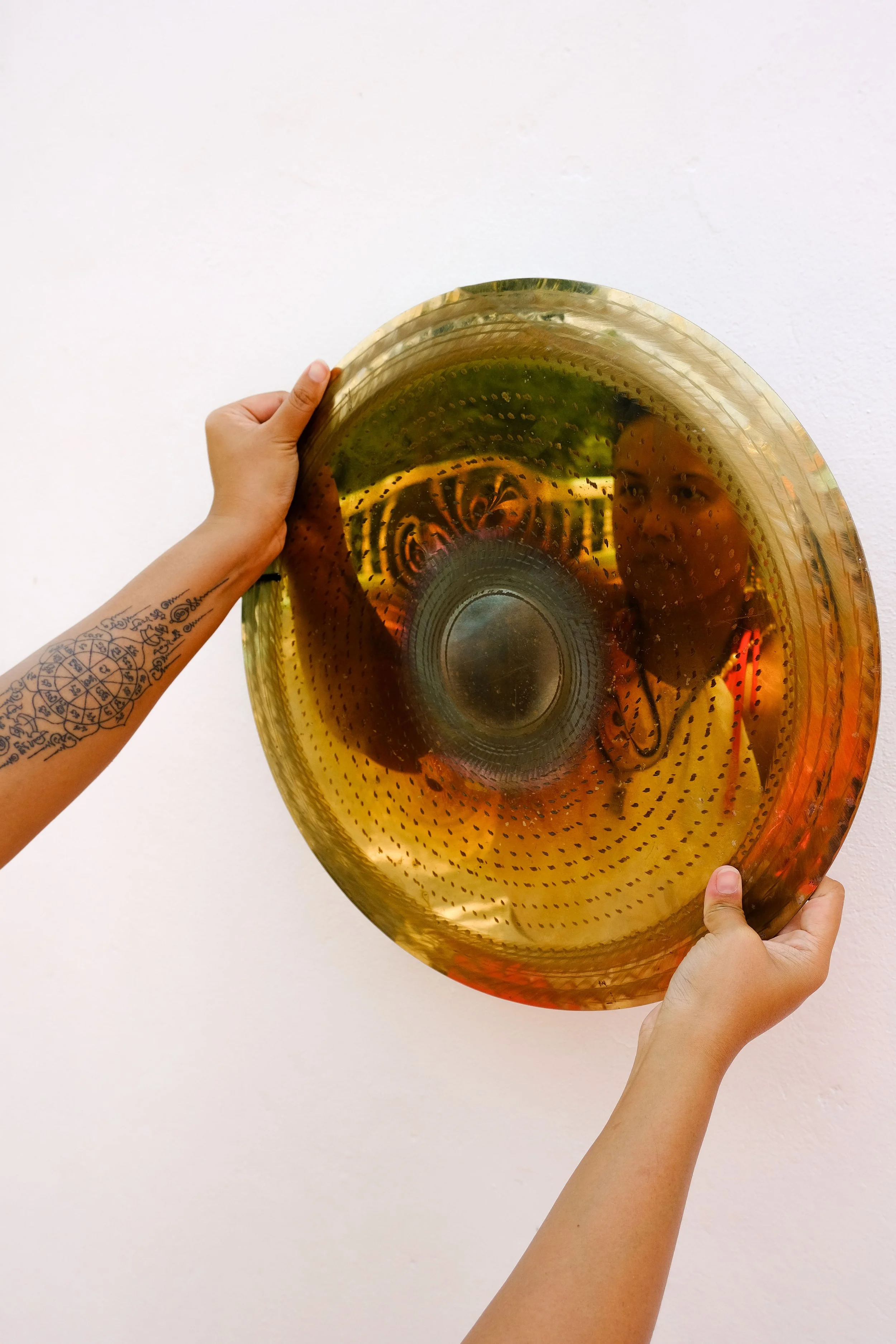 Person holding a large, gold-colored, perforated glass dish reflecting their face and arm tattoos, with a plain white wall in the background.