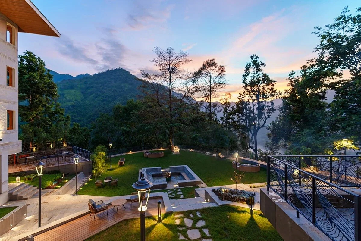 An outdoor terrace area at dusk, with a mountain view, surrounded by trees, with seating and lighting.