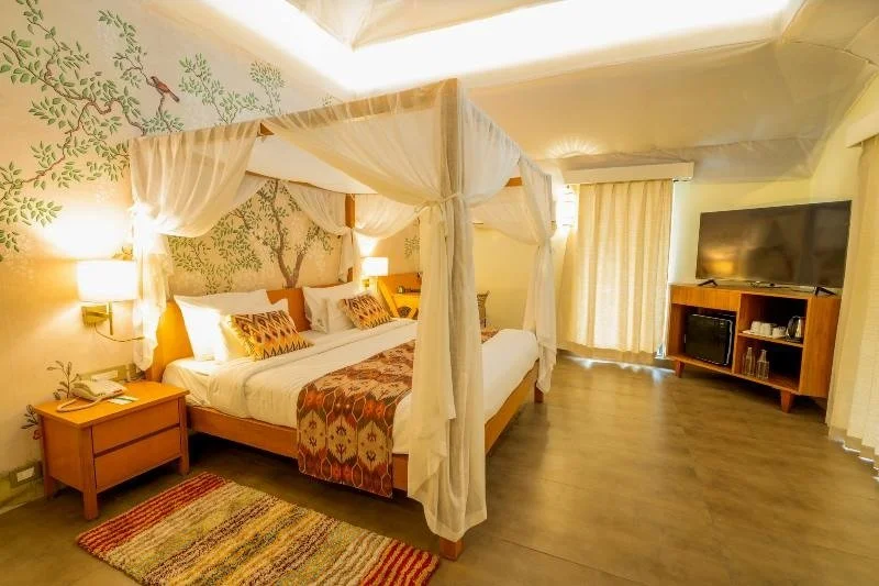 Room with four-poster bed, decorative pillows, wall mural, TV, dresser, lamp, and window with curtains.