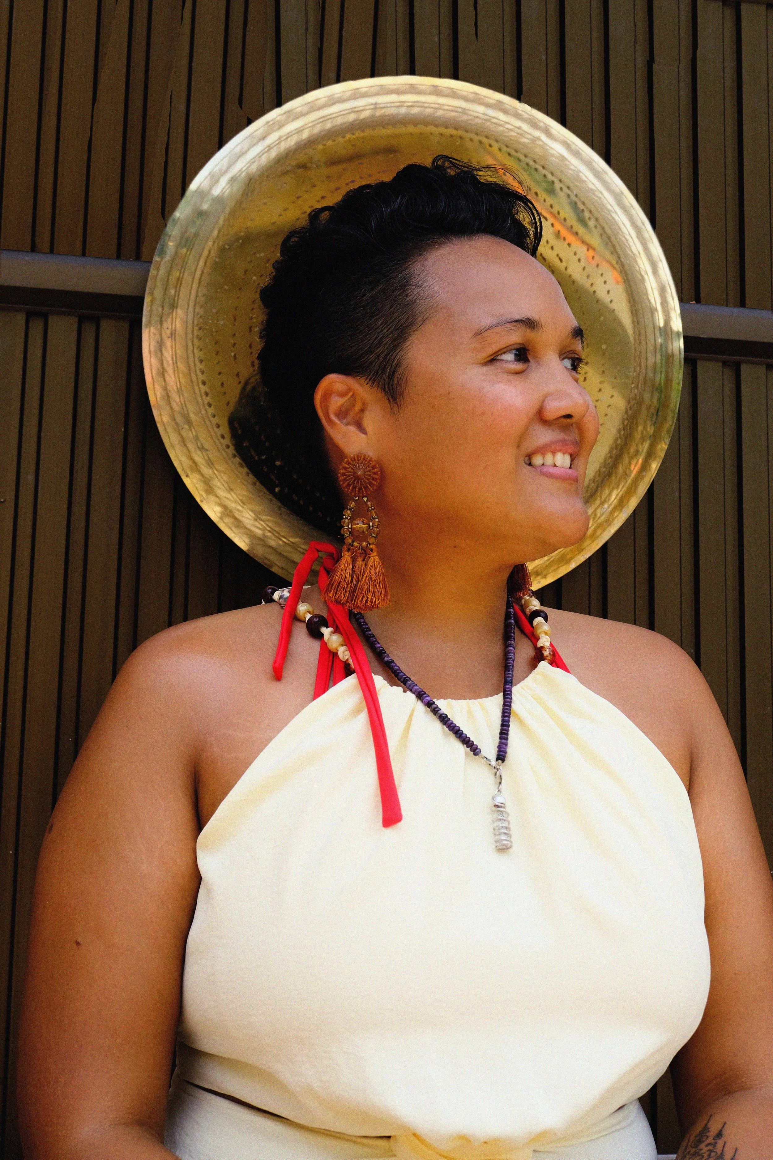 Woman wearing a large straw hat, colorful earrings, necklace, and a white dress, smiling and looking to the side.