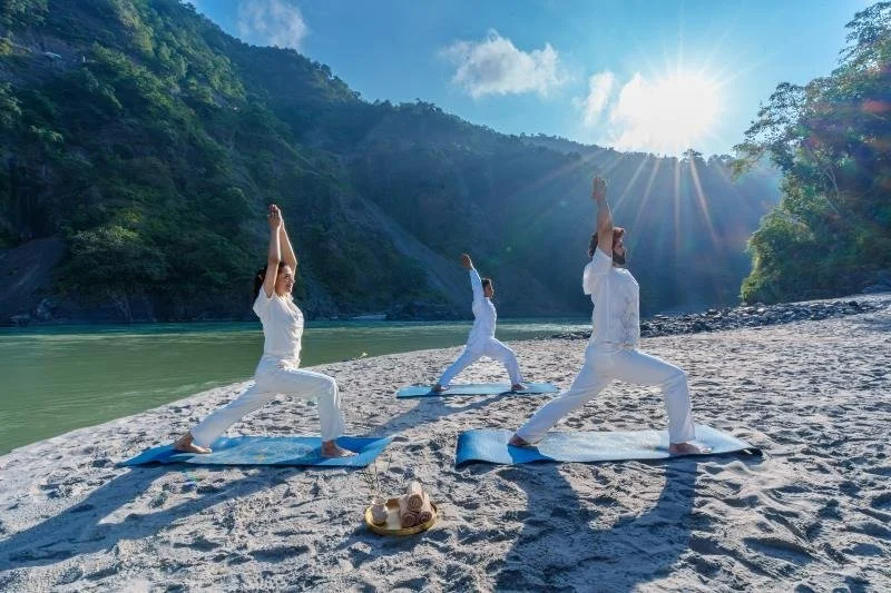 Three people practicing yoga on mats on a sandy beach by a river, with green mountains and a bright sun in the sky.