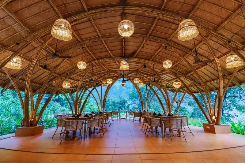 Open-air restaurant with bamboo-arched ceiling, hanging woven lamps, and dining tables with chairs, overlooking a lush green landscape.