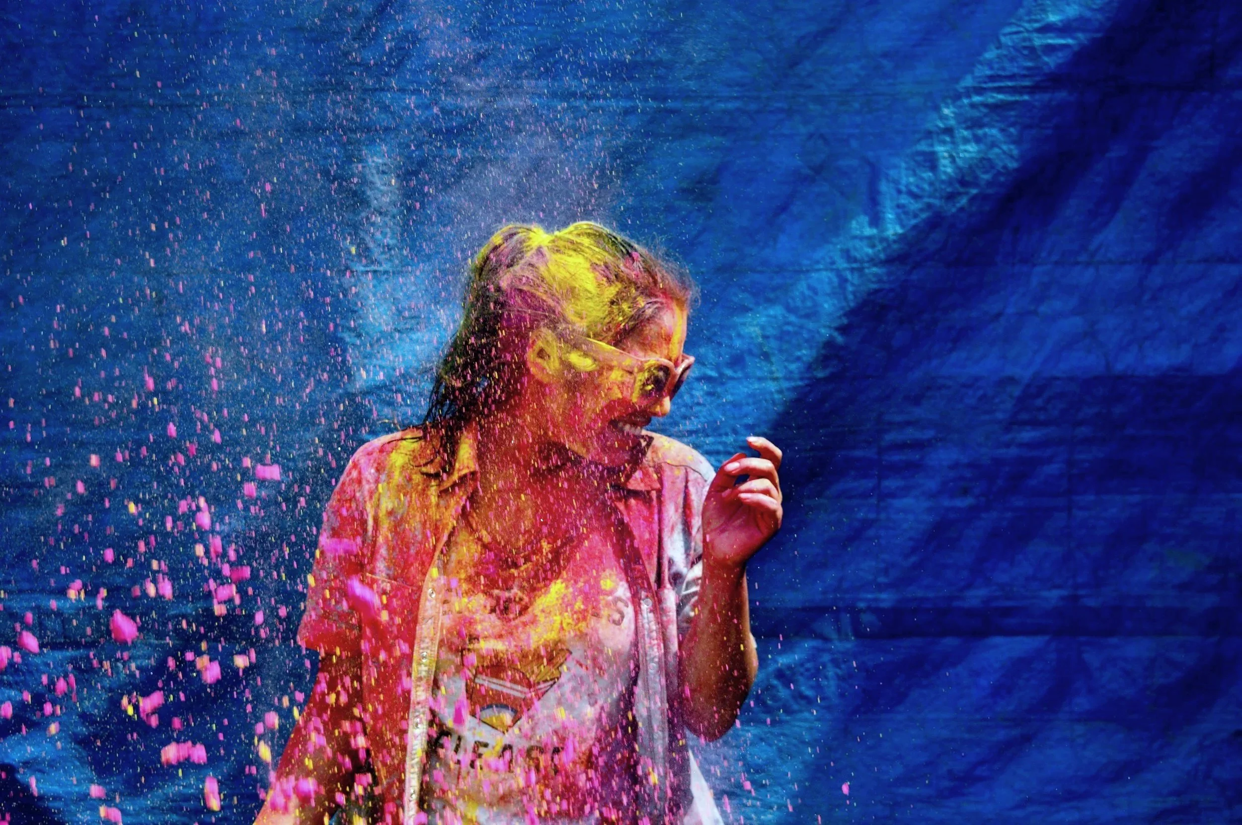 A woman covered in pink, yellow, and purple powder, wearing glasses and a light-colored shirt, appears to be celebrating during a colorful event with a blue background.