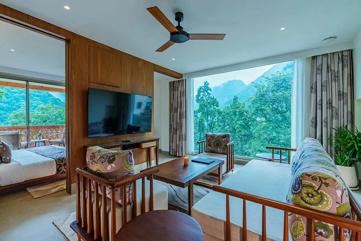 A bright living room with large windows showing a lush green mountain landscape. Contains a sofa, two armchairs with colorful floral cushions, a wooden coffee table, and a flat-screen TV mounted on a wooden-paneled wall. In the background, a bedroom with a bed and sliding glass doors to a balcony.