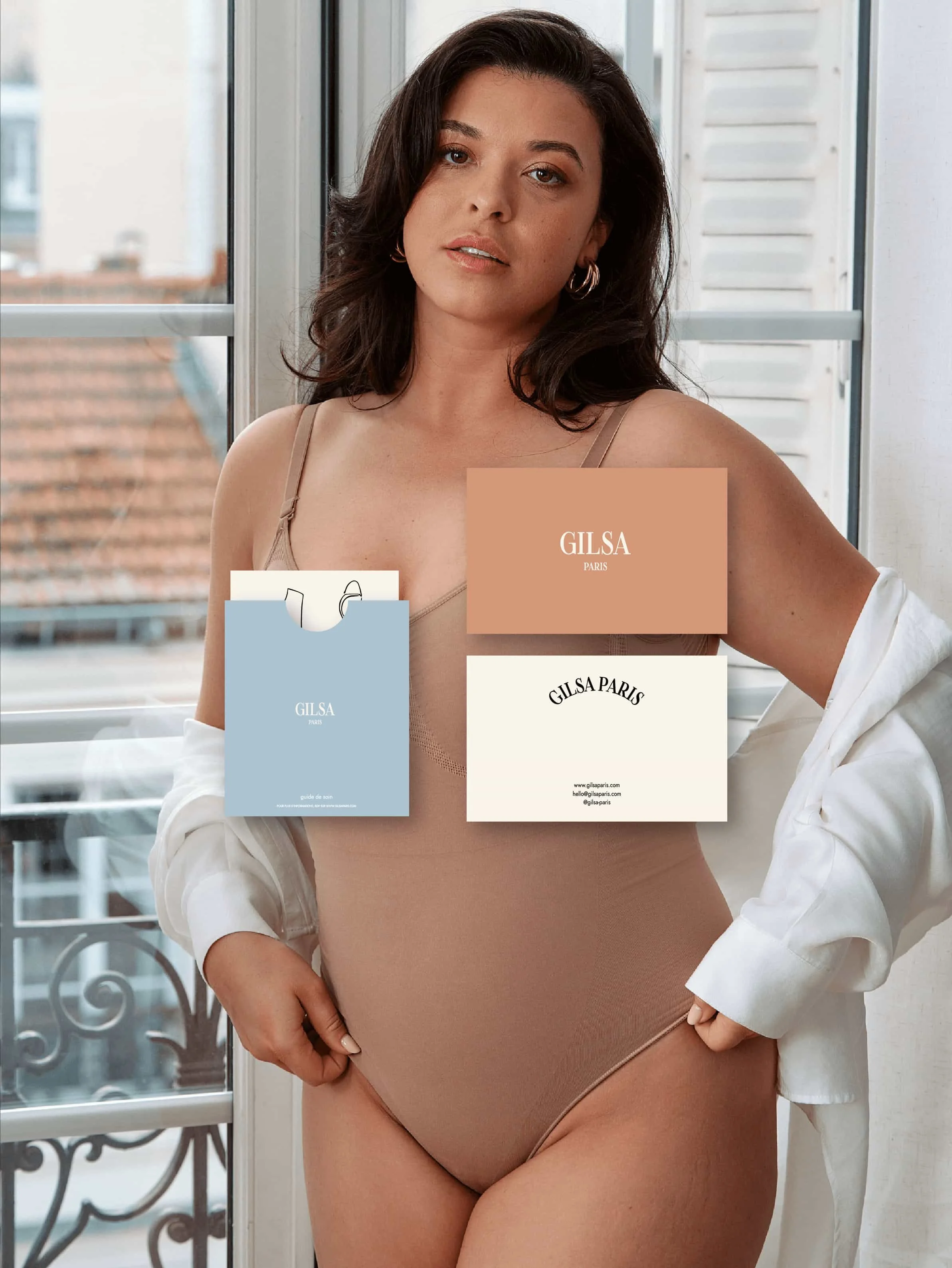 Une jeune femme brune porte un body beige en coton, debout près d'une fenêtre avec un cadre en bois blanc. Elle porte des boucles d'oreilles en forme de cercles et regarde l'appareil photo avec une expression neutre.