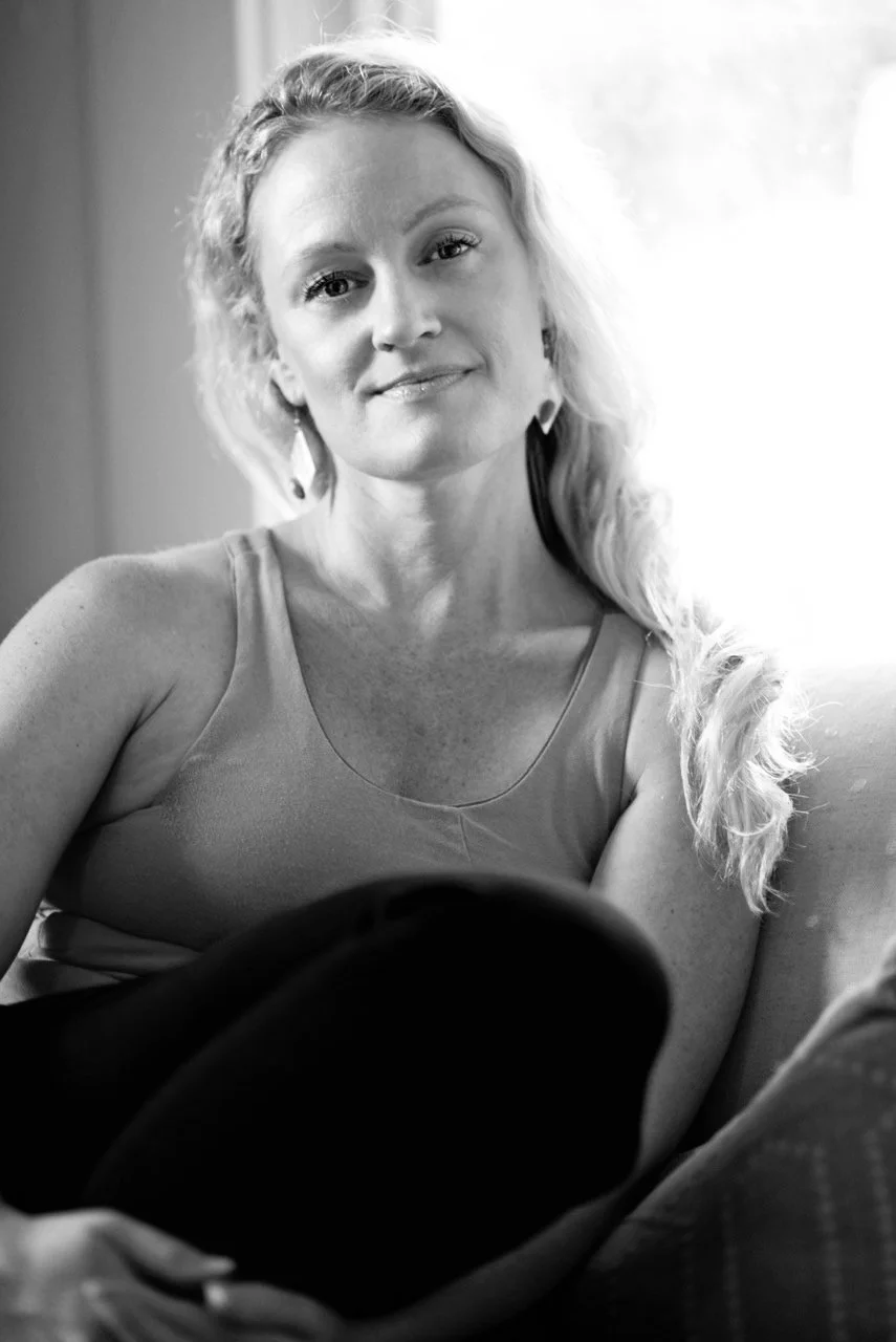 A woman with long, wavy hair sitting on a couch, smiling gently, with a window in the background.
