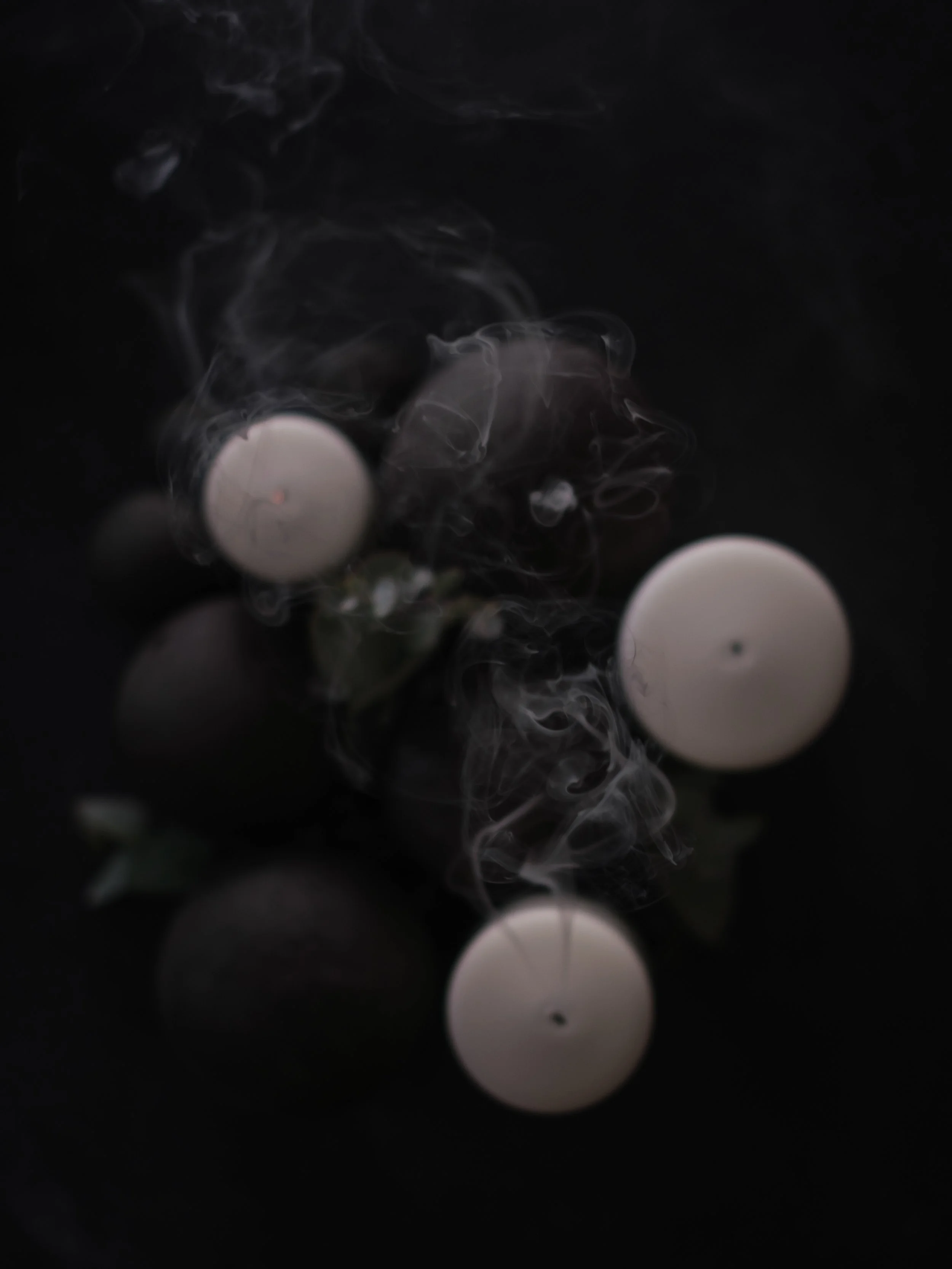 White candles surrounded by dark ceramics and smoke against a black background.