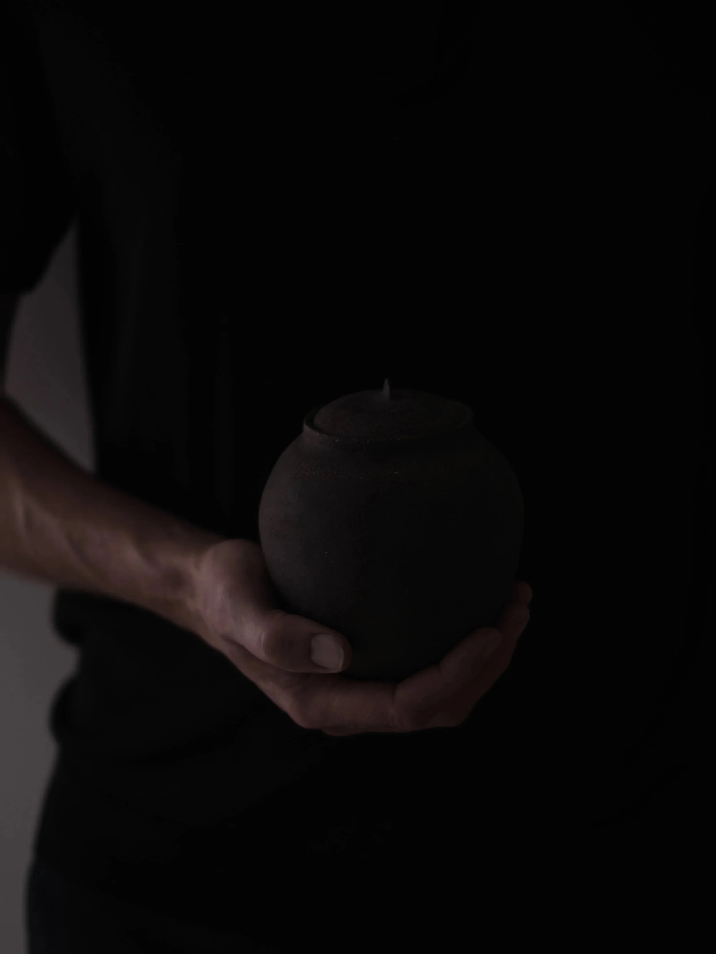 Person holding a dark, rounded ceramic pot in a dimly lit environment.