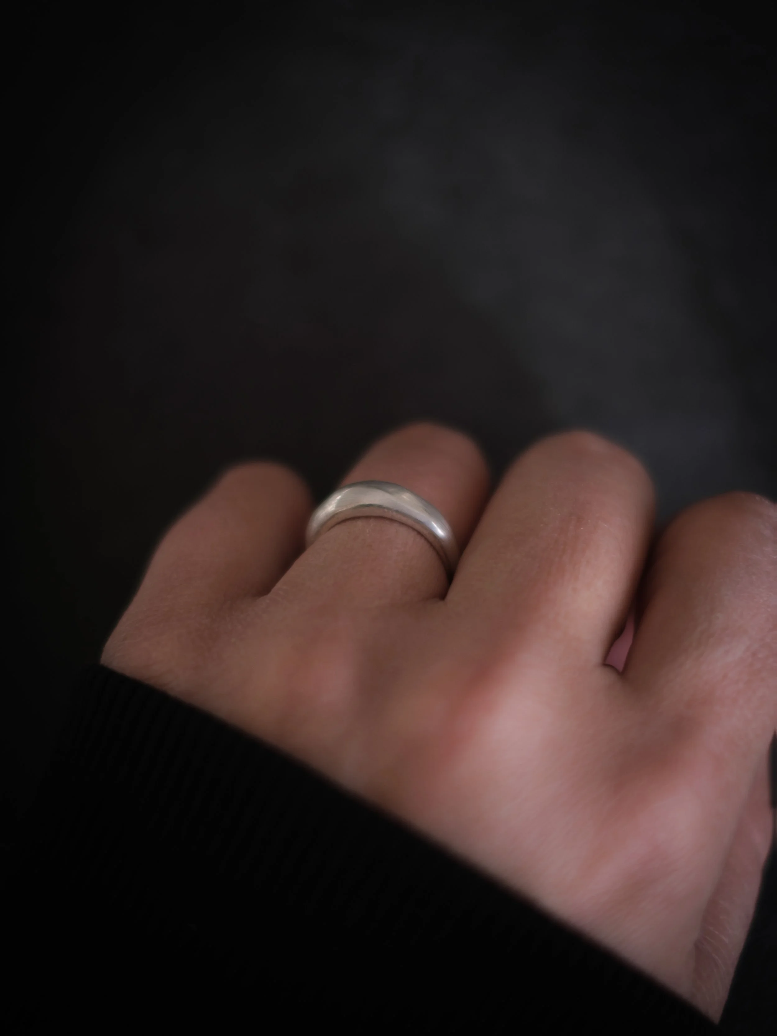 Close-up of a hand wearing a silver ring on the middle finger, with a black sleeve on the wrist, against a dark background.
