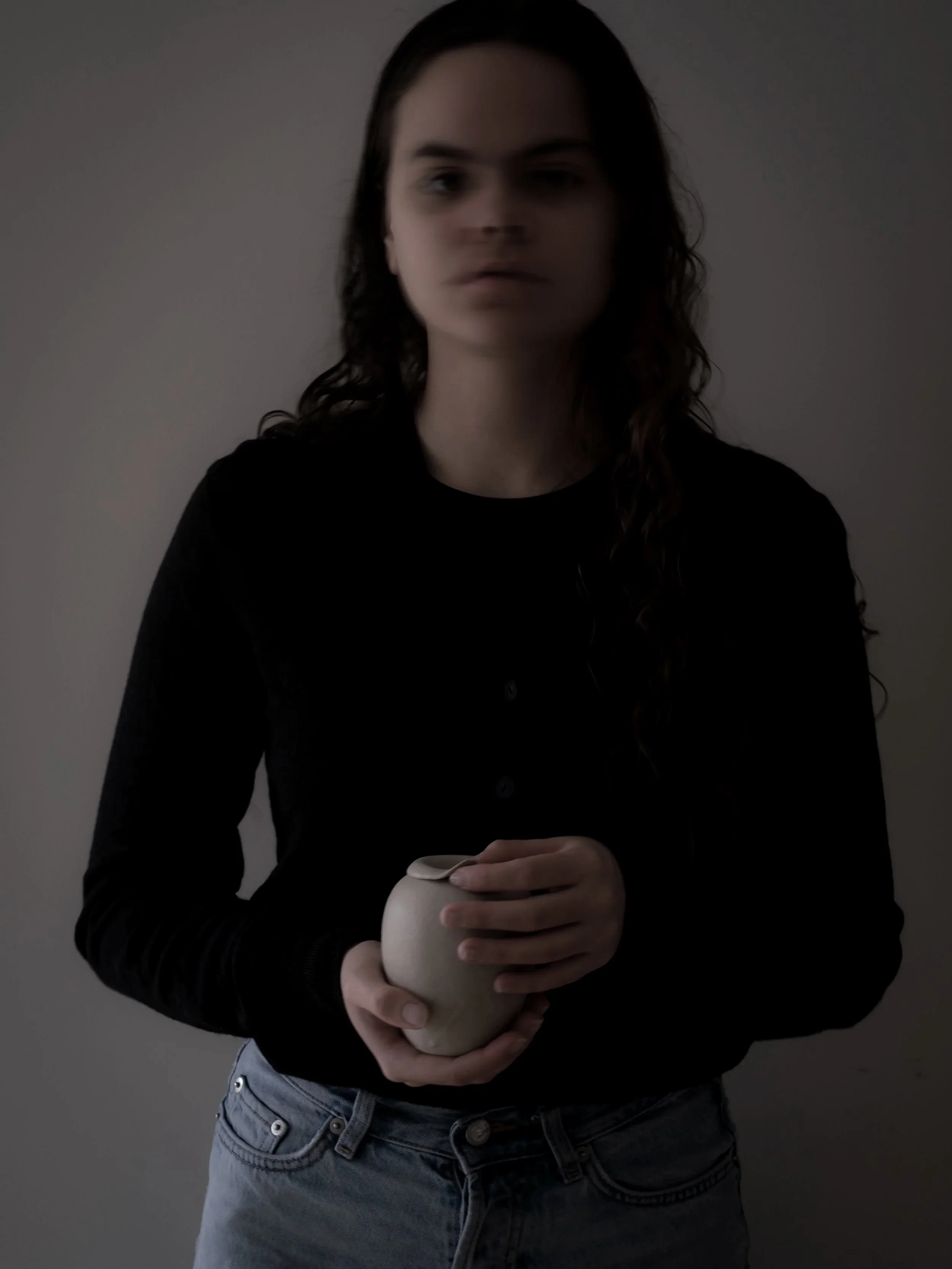 A woman with long dark hair holding a ceramic urn, standing against a plain background.