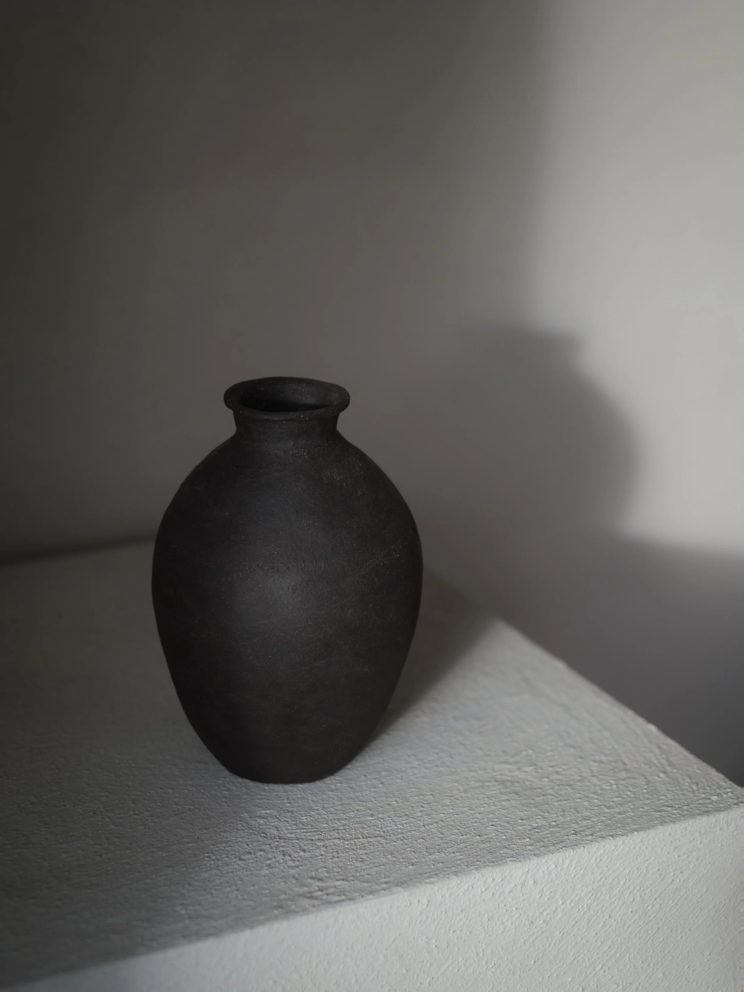 A dark, textured ceramic vase on a white pedestal with dramatic lighting and shadows.