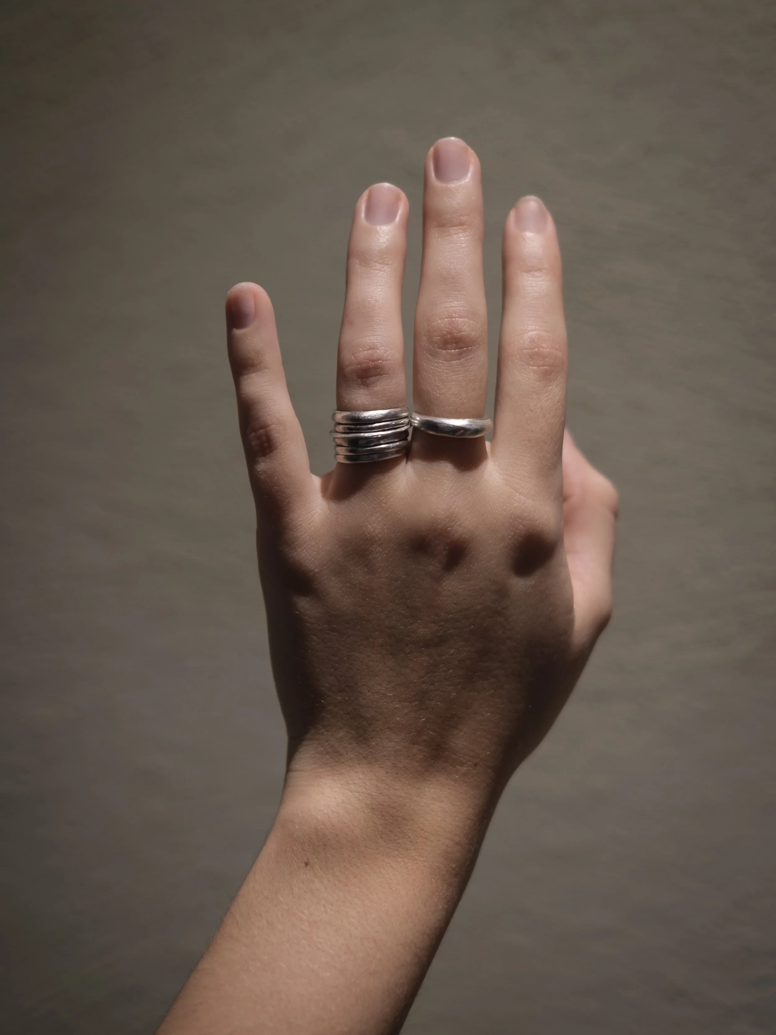 A person's hand with fingers raised, wearing multiple silver rings, against a plain background.