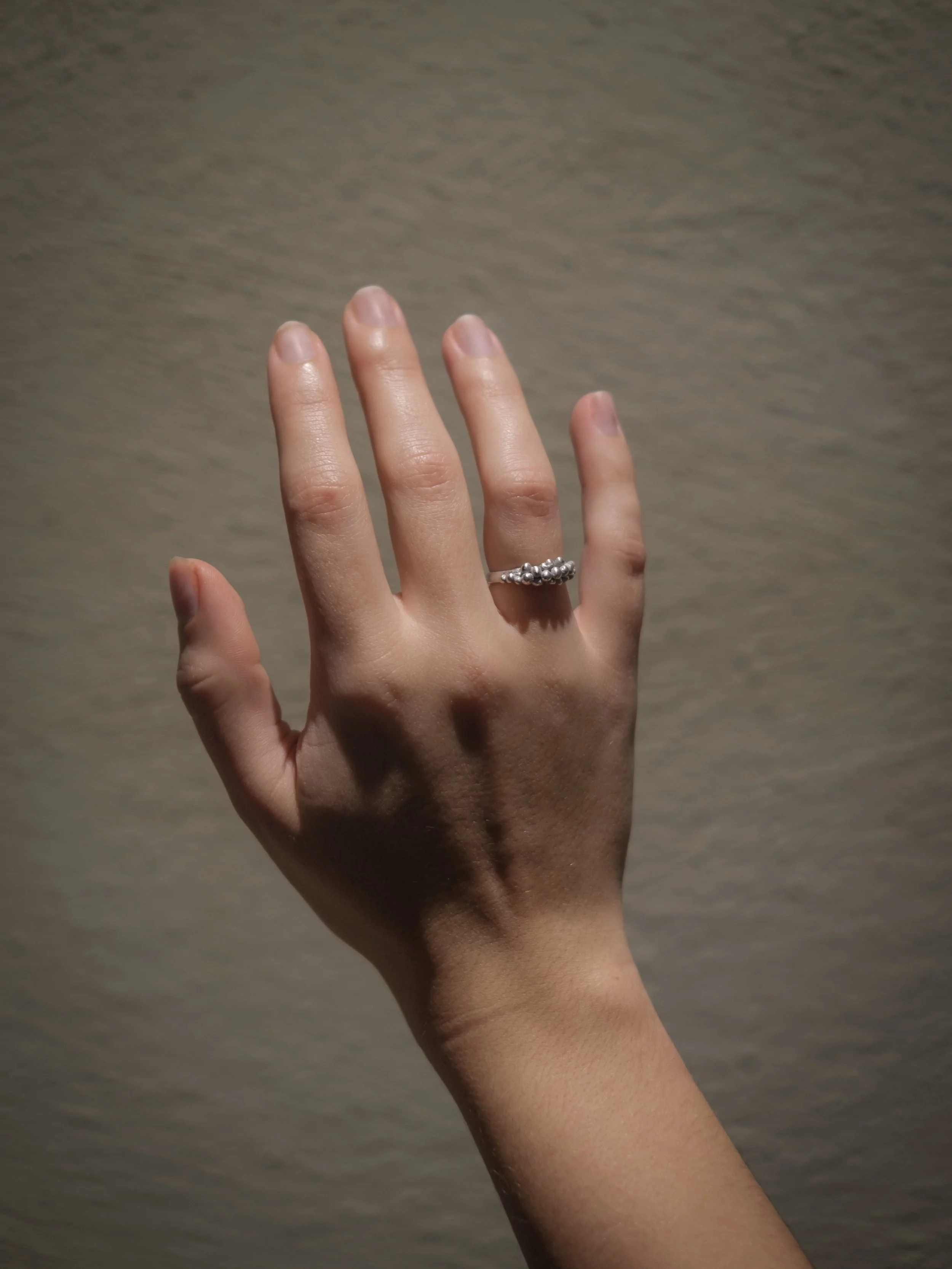 A hand with fair skin and short, neatly trimmed nails is raised against a plain beige background. The hand is wearing a silver ring with small decorative beads on the ring finger.