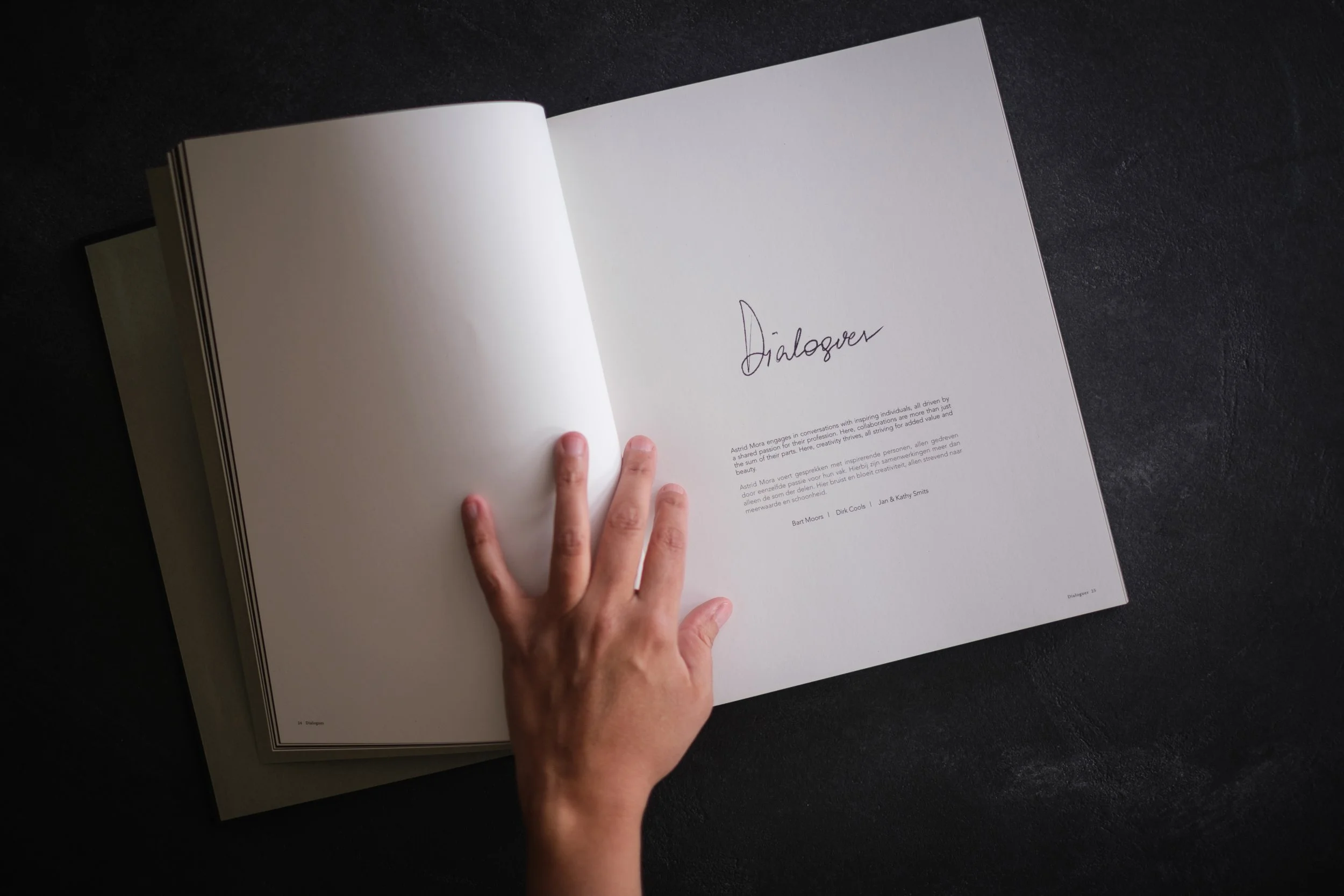 Open book on a dark surface with a person's hand turning the page. The right page has the word 'Dialogues' written, with smaller text below.
