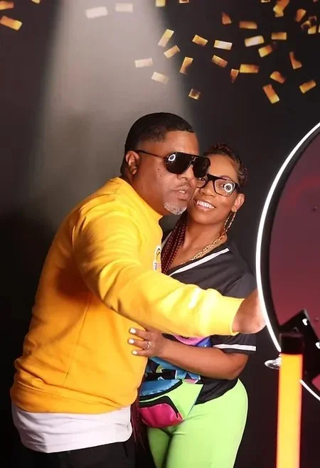 A smiling couple posing for a photo booth with confetti falling in the background. The man is wearing a yellow sweater and sunglasses, and the woman is wearing glasses and colorful clothing. They appear to be enjoying a festive event.