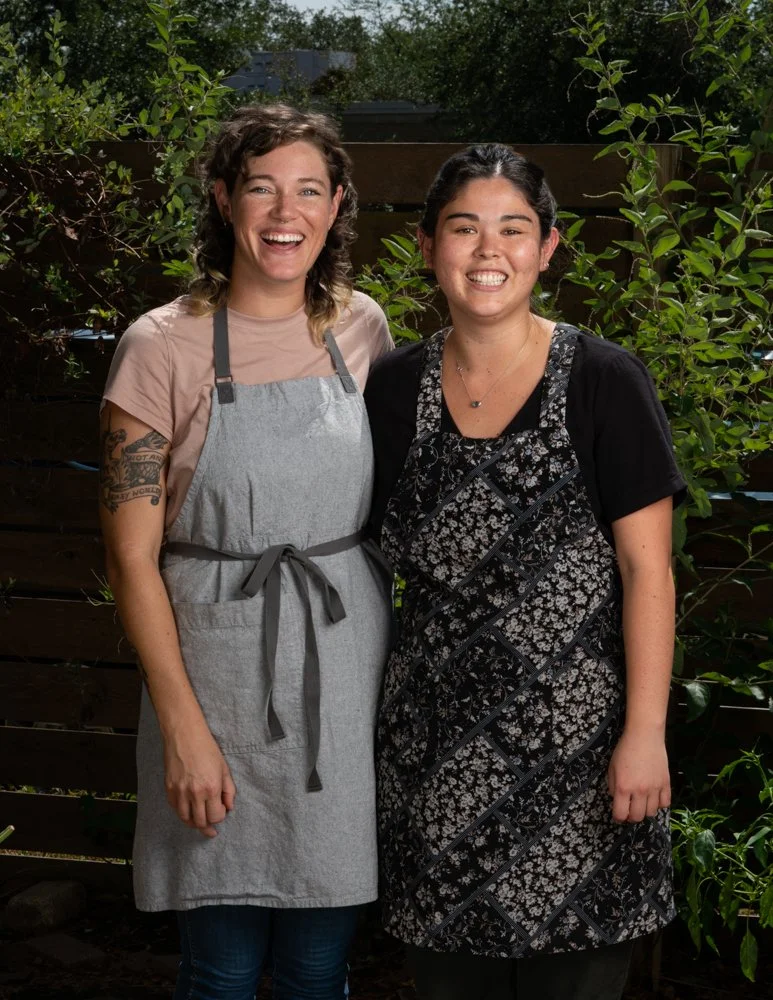Two women outdoors dressed casually wearing aprons smiling together for this editorial style photo.