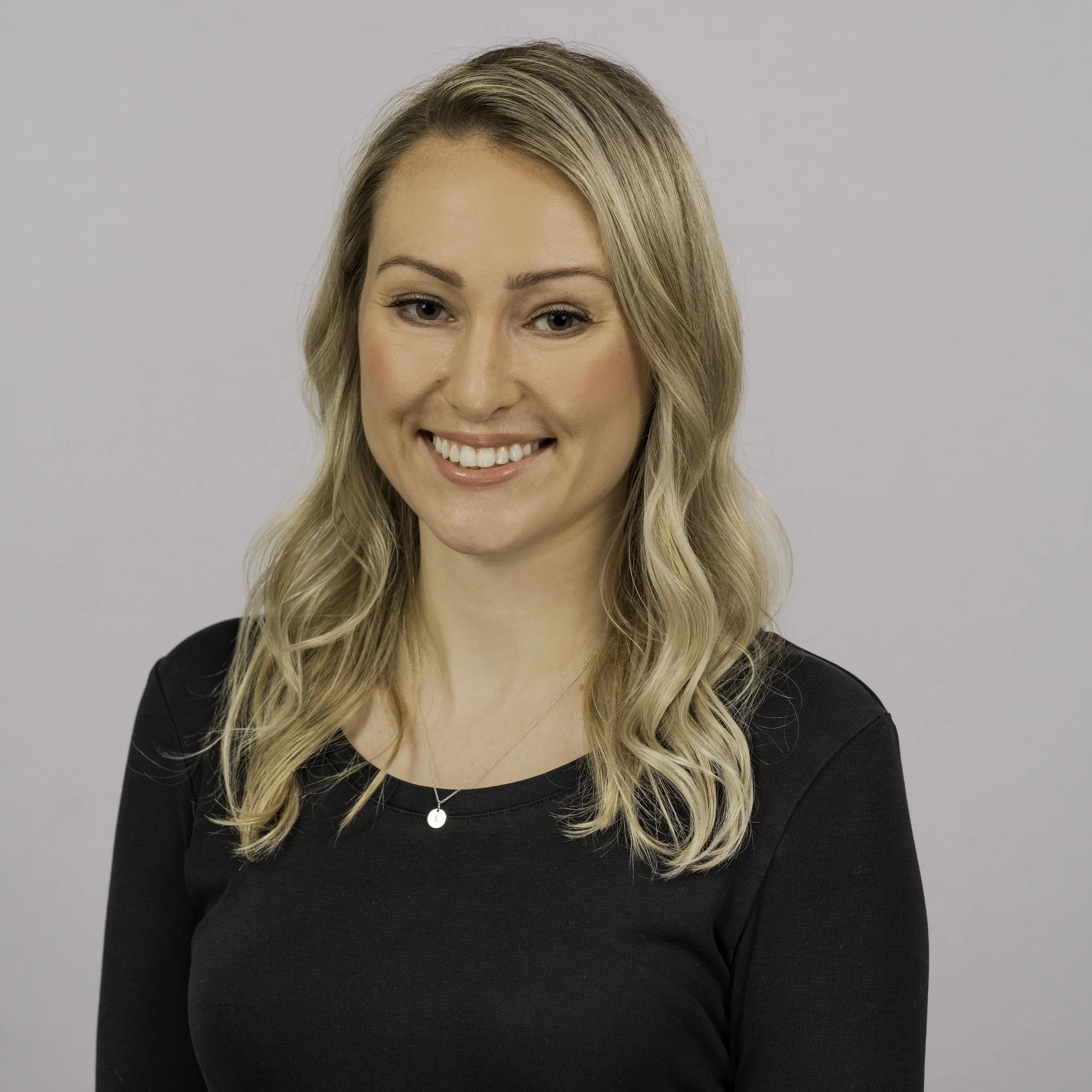 A woman with blonde, wavy hair and gray eyes smiling in front of a plain light gray background. She is wearing a black top and layered necklaces.
