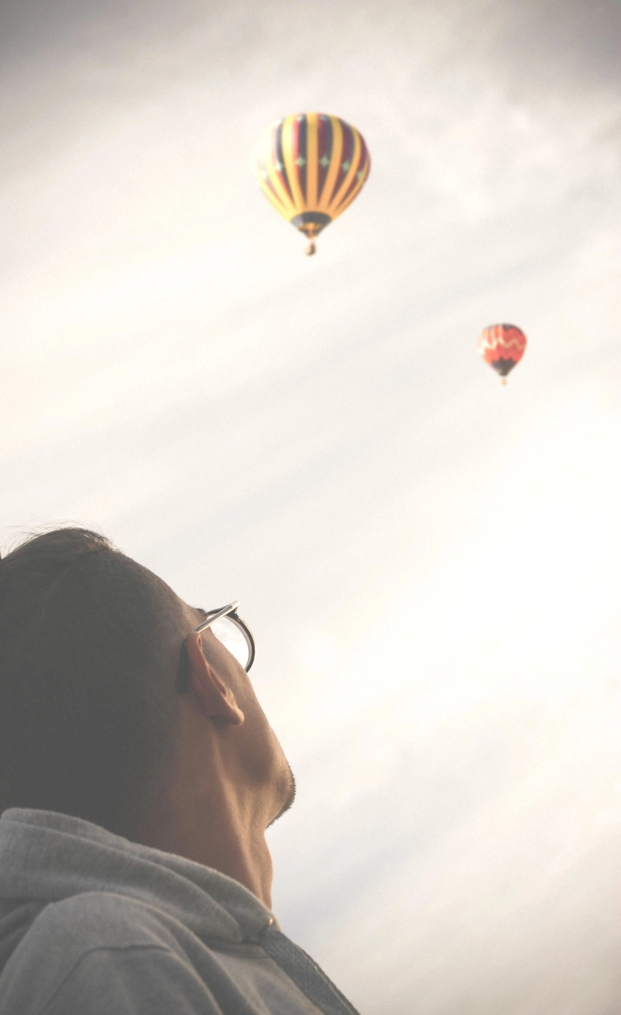 A man with glasses is looking up at two hot air balloons floating in the sky.