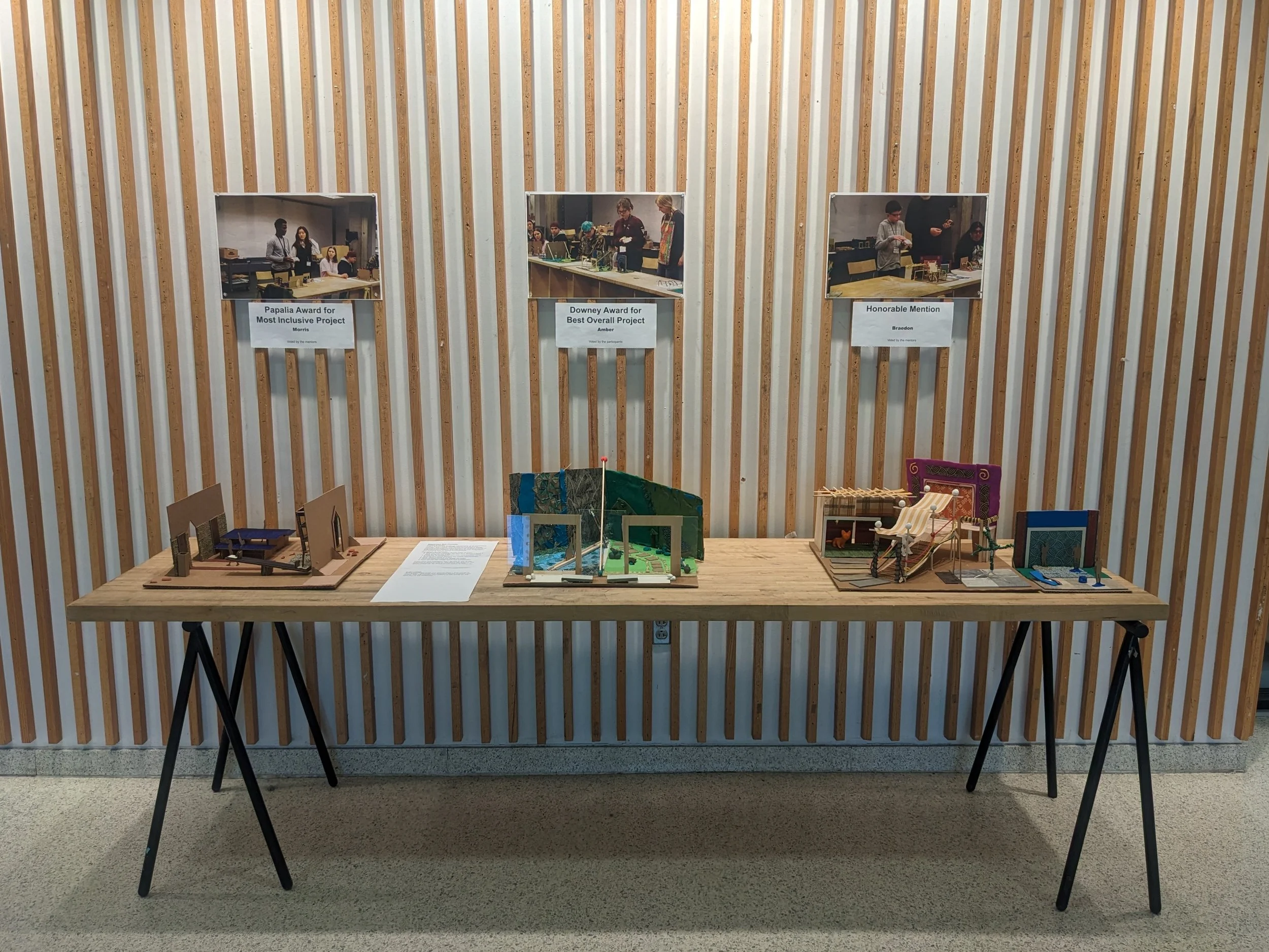 Participants' final models exhibited in the lobby of Cowgill Hall. ID: Three models sit on a butcher block table against a wood slat wall. Pictures of the participants are pinned above each model.