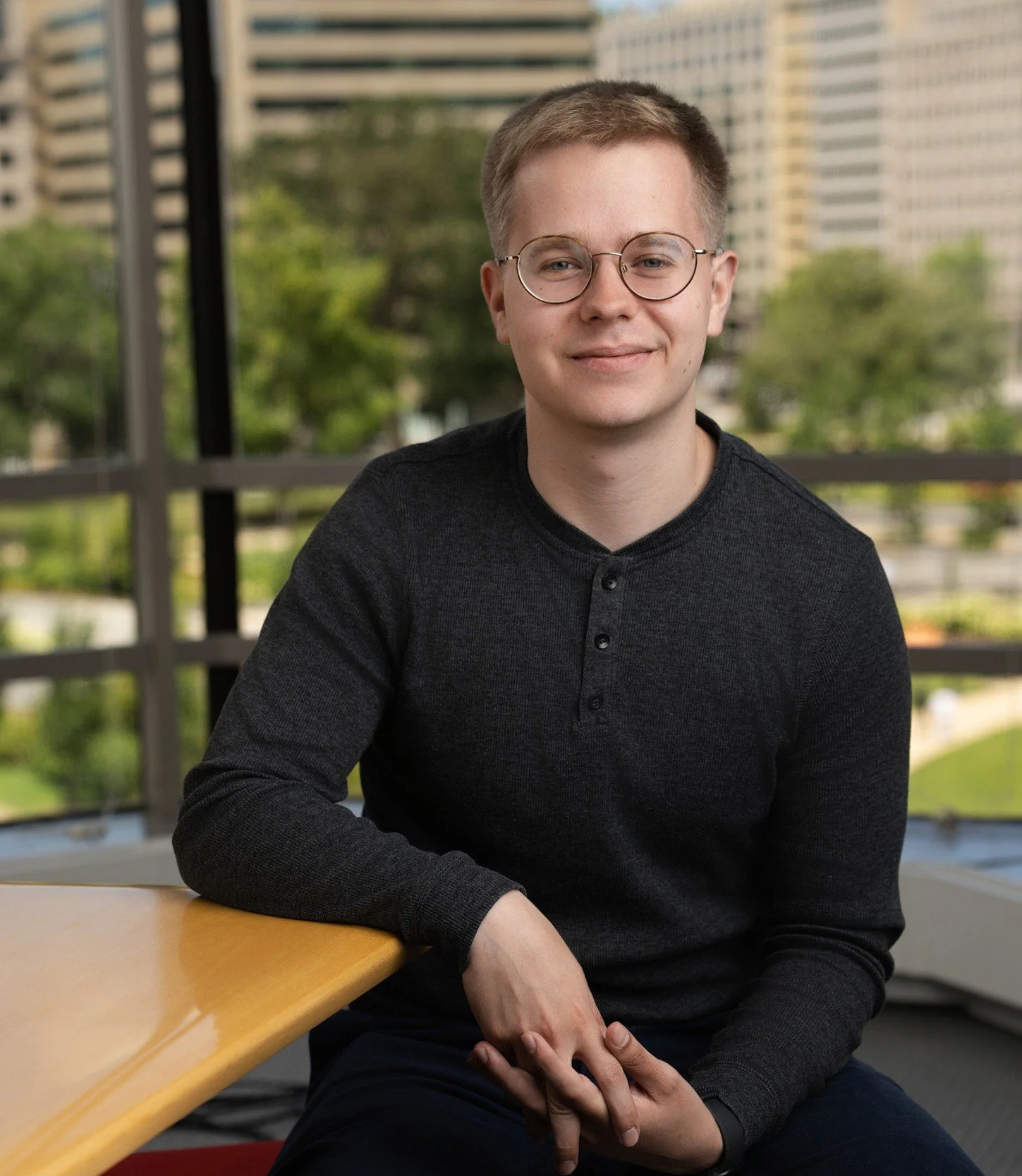 Portrait of a young white man with short blond hair, round glasses, and a black henley shirt, sitting with one arm resting on a wood table.